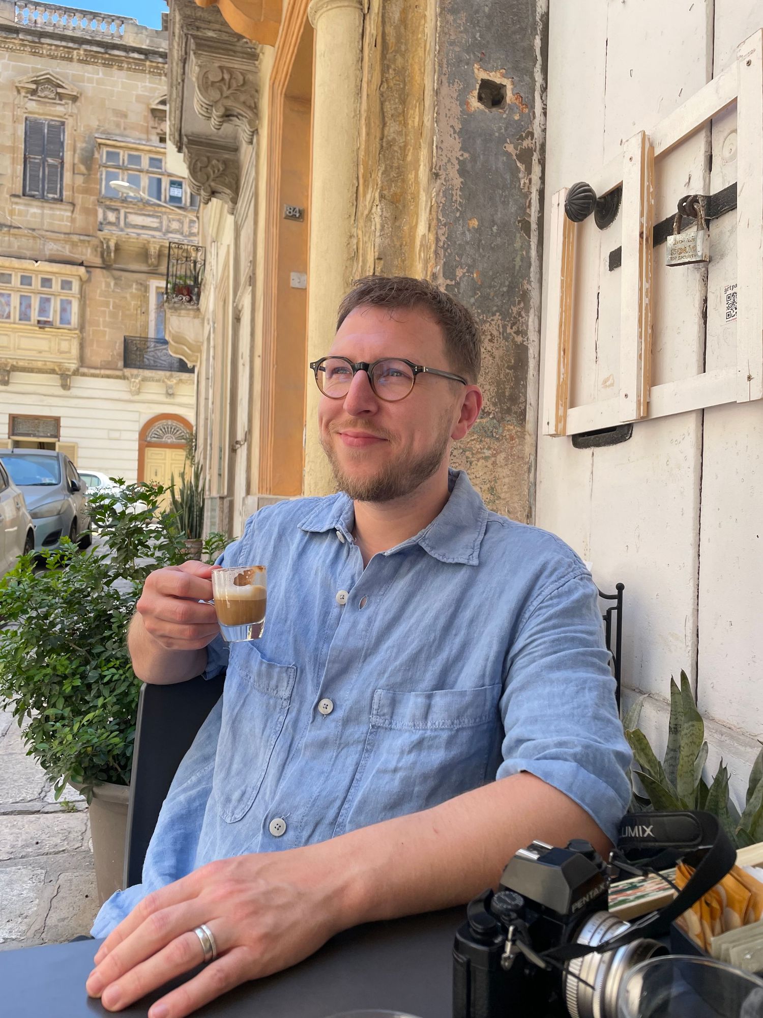 Person in glasses and blue shirt enjoying a drink at an outdoor cafe with historic European architecture in background.