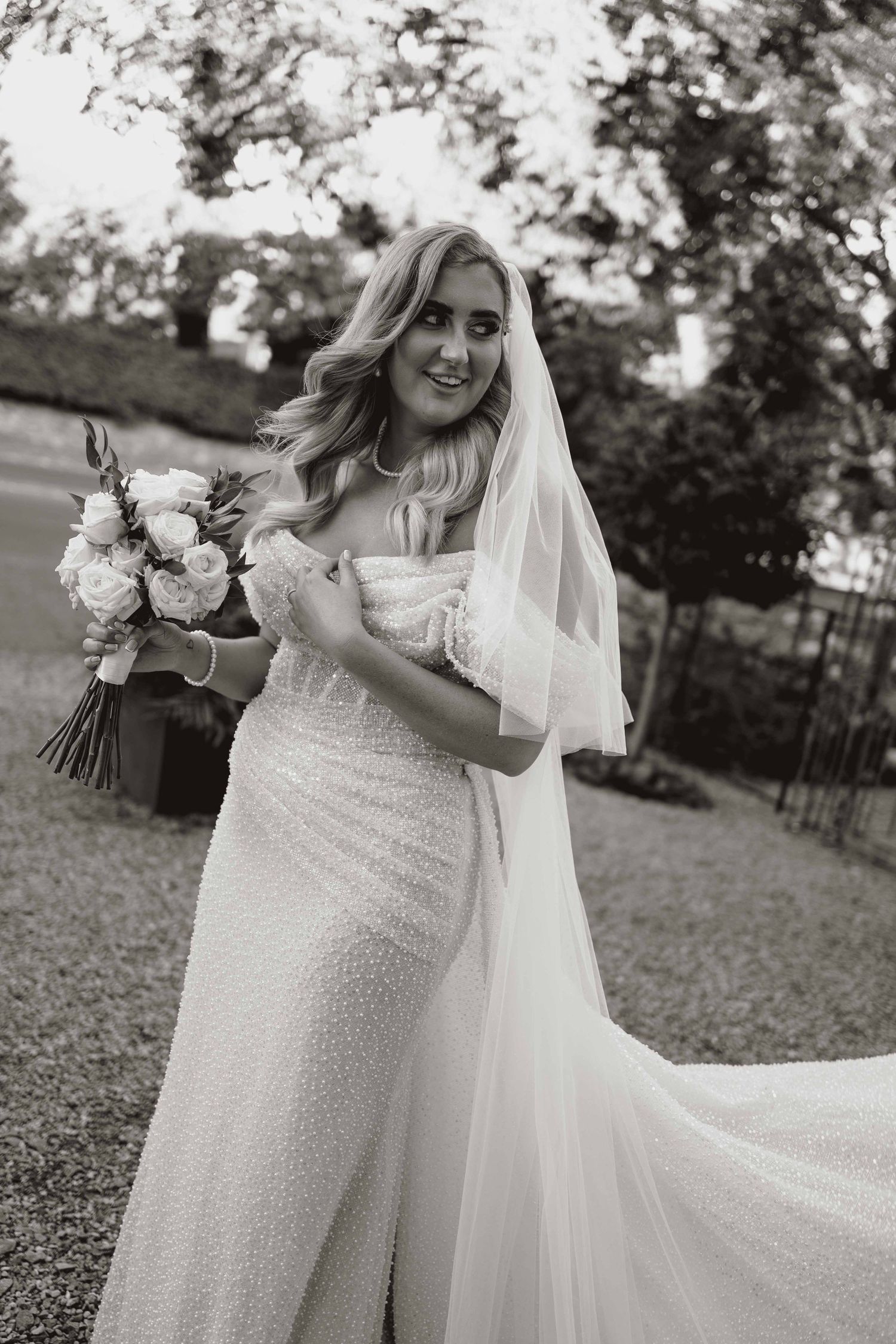A bride in a sequined wedding dress poses joyfully outdoors while holding a white floral bouquet.
