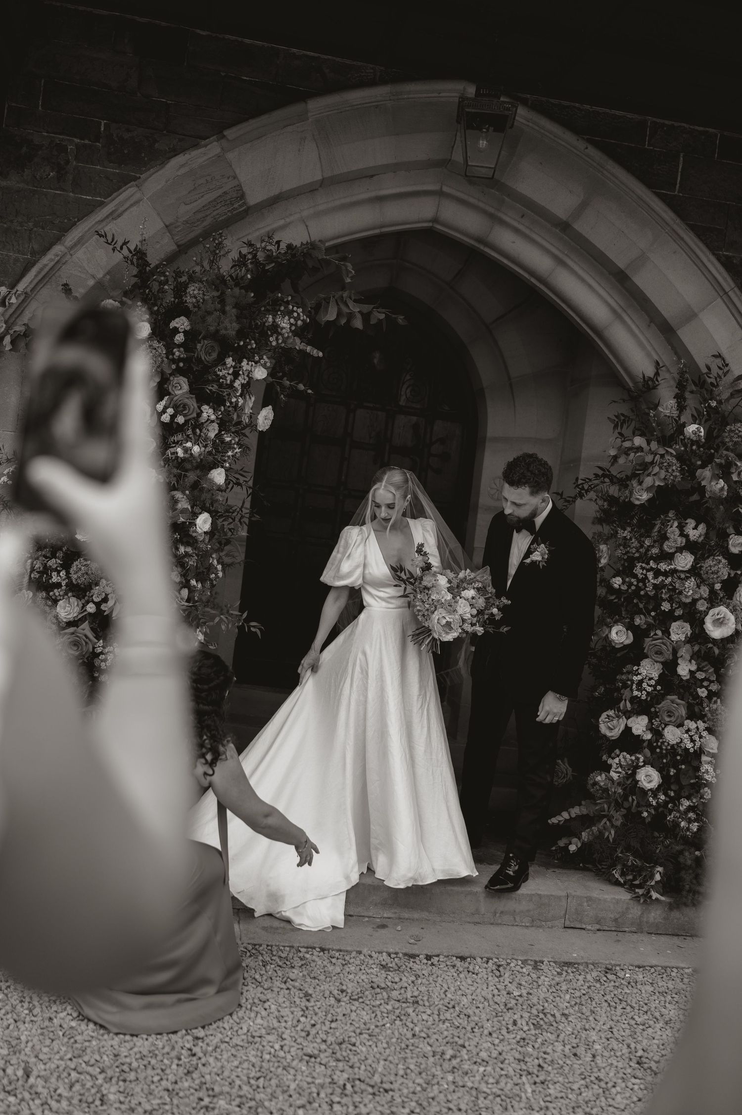 Black and white wedding photos show a couple twirling and dancing together under a stone archway decorated with flowers.