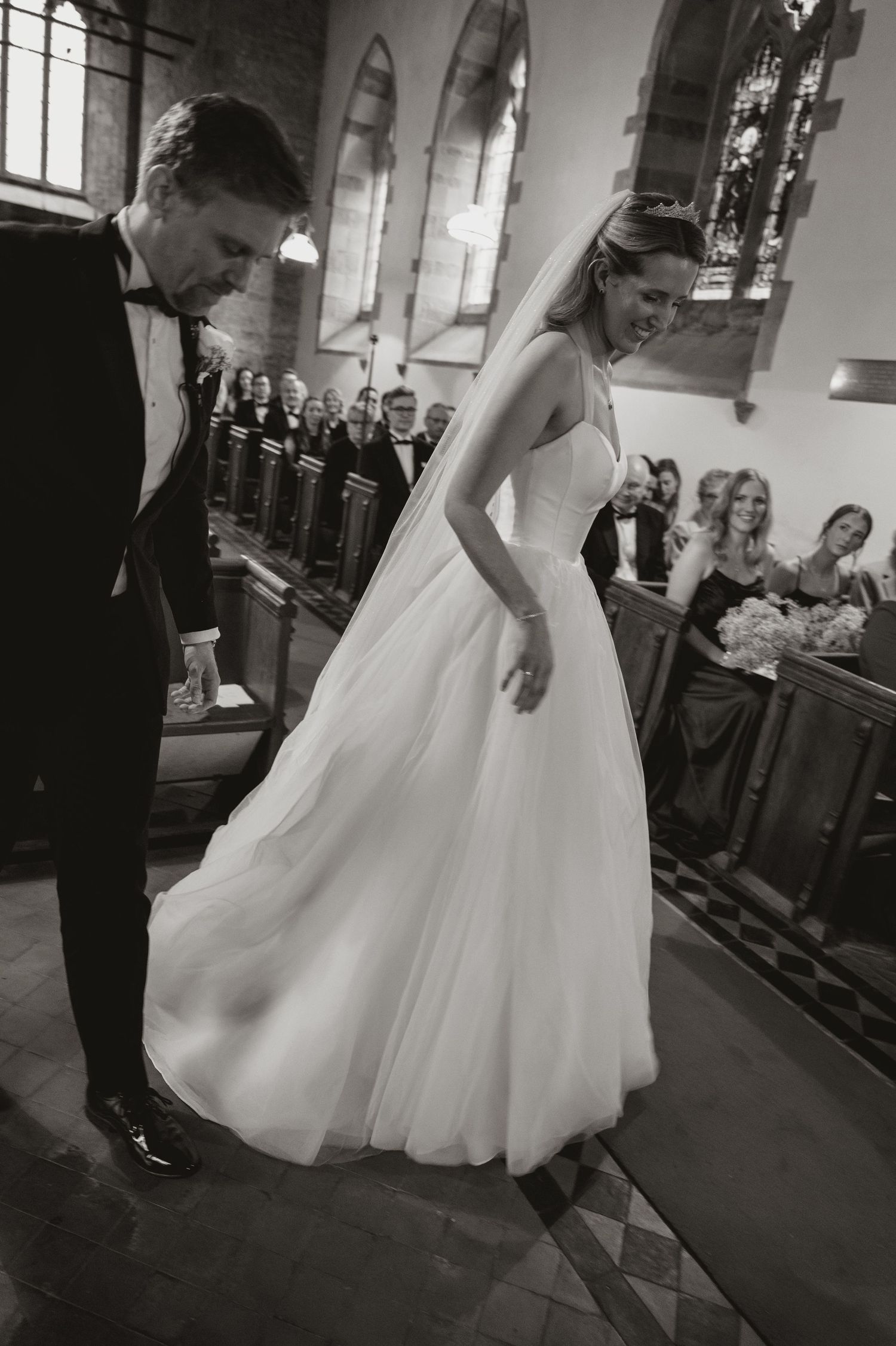 A black and white wedding photograph showing a couple in formal attire inside a church with stained glass windows.