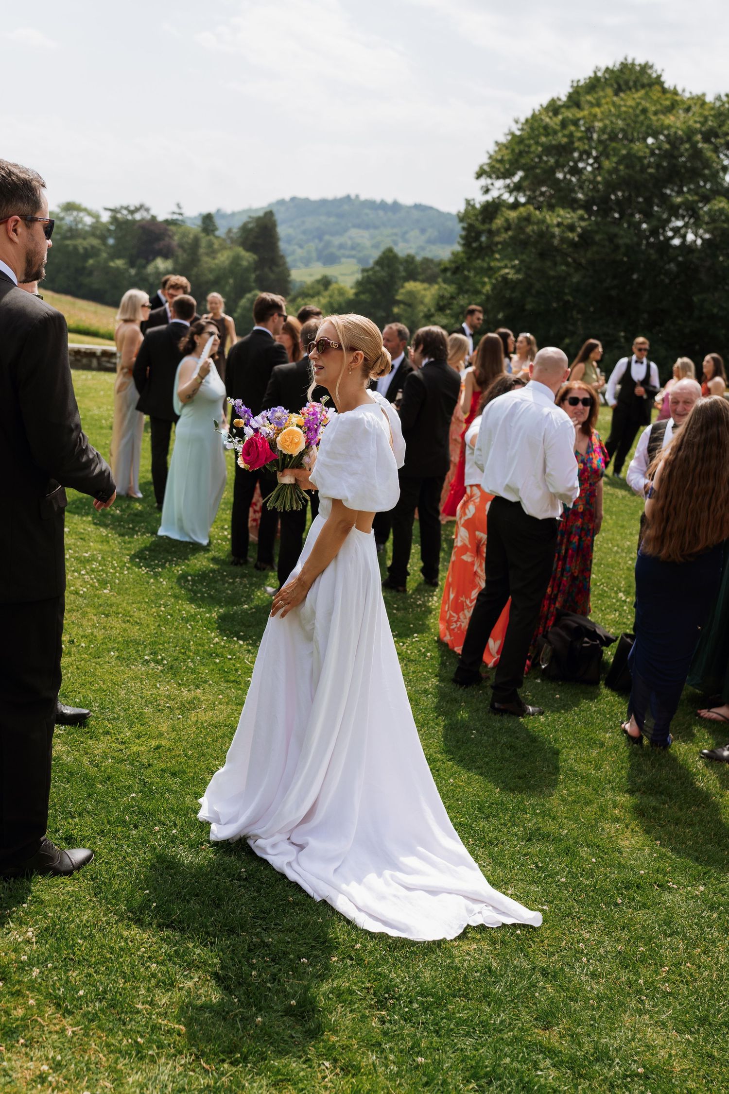 Wedding guests gather on a sunny hillside lawn as a couple in white shares a romantic embrace during an outdoor ceremony.