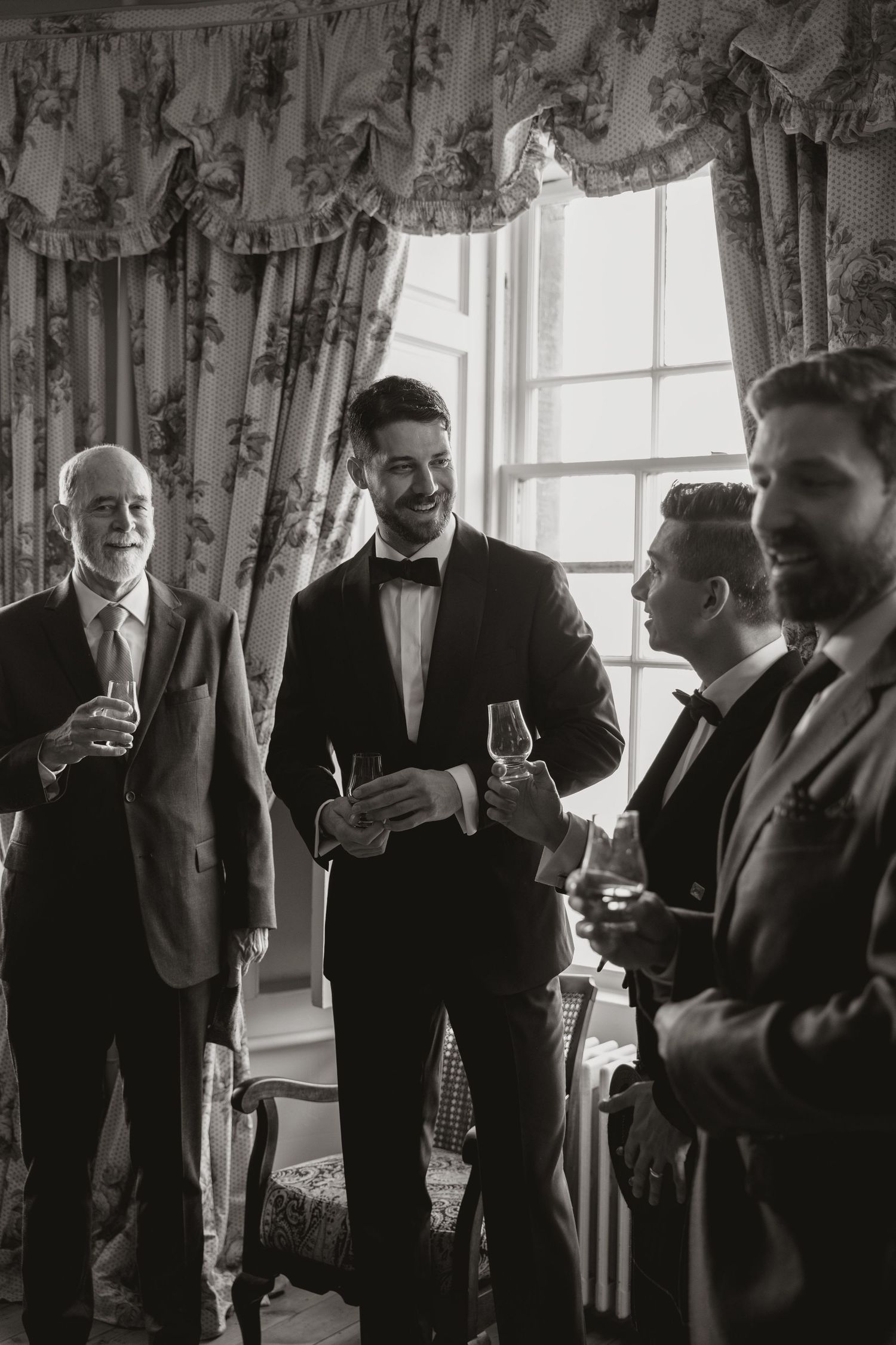 A group of people in formal attire having drinks and conversation by a window with elegant curtains in black and white.