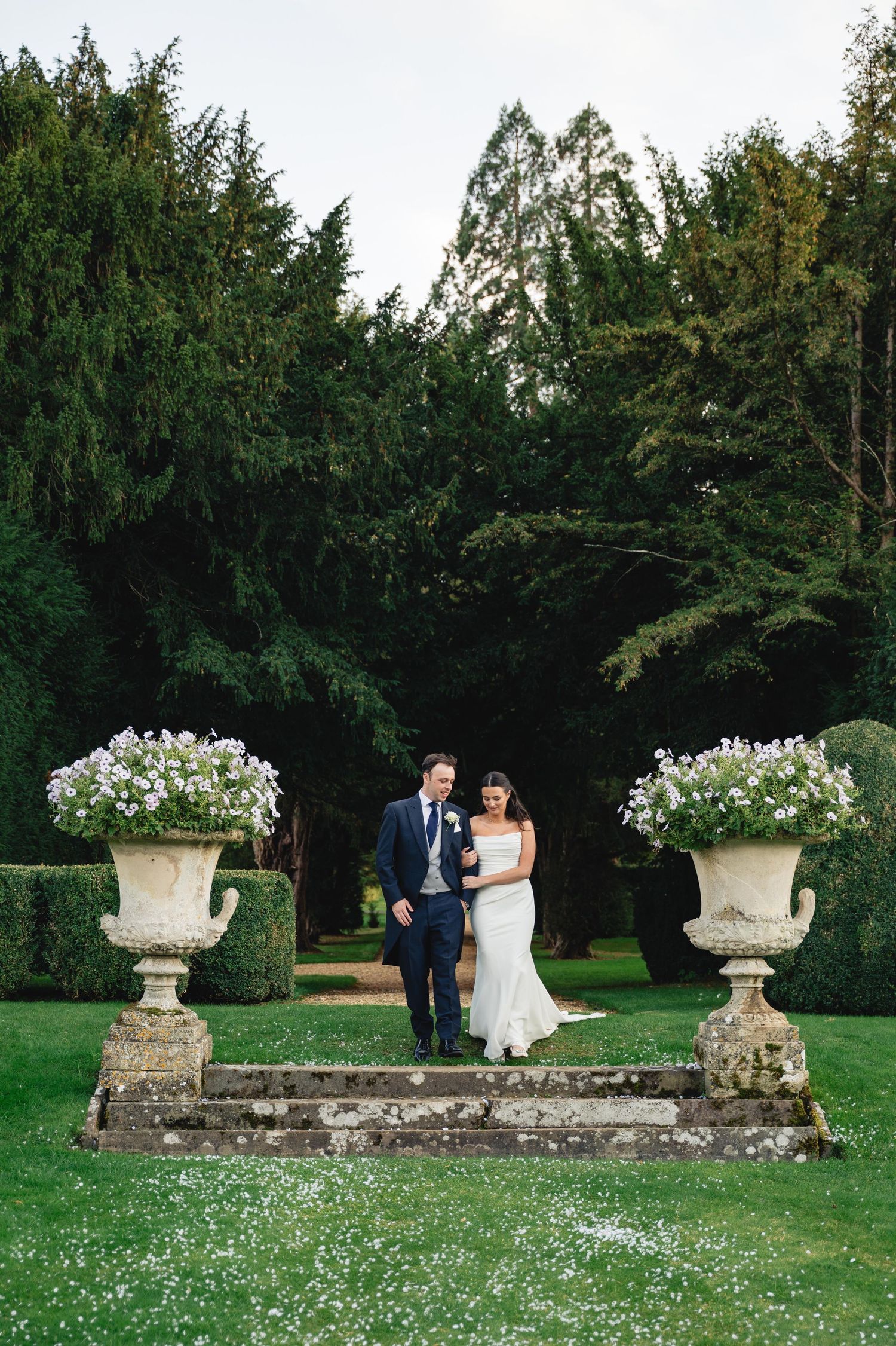 A couple poses near ornate stone garden urns with white flowers in a lush green garden setting.