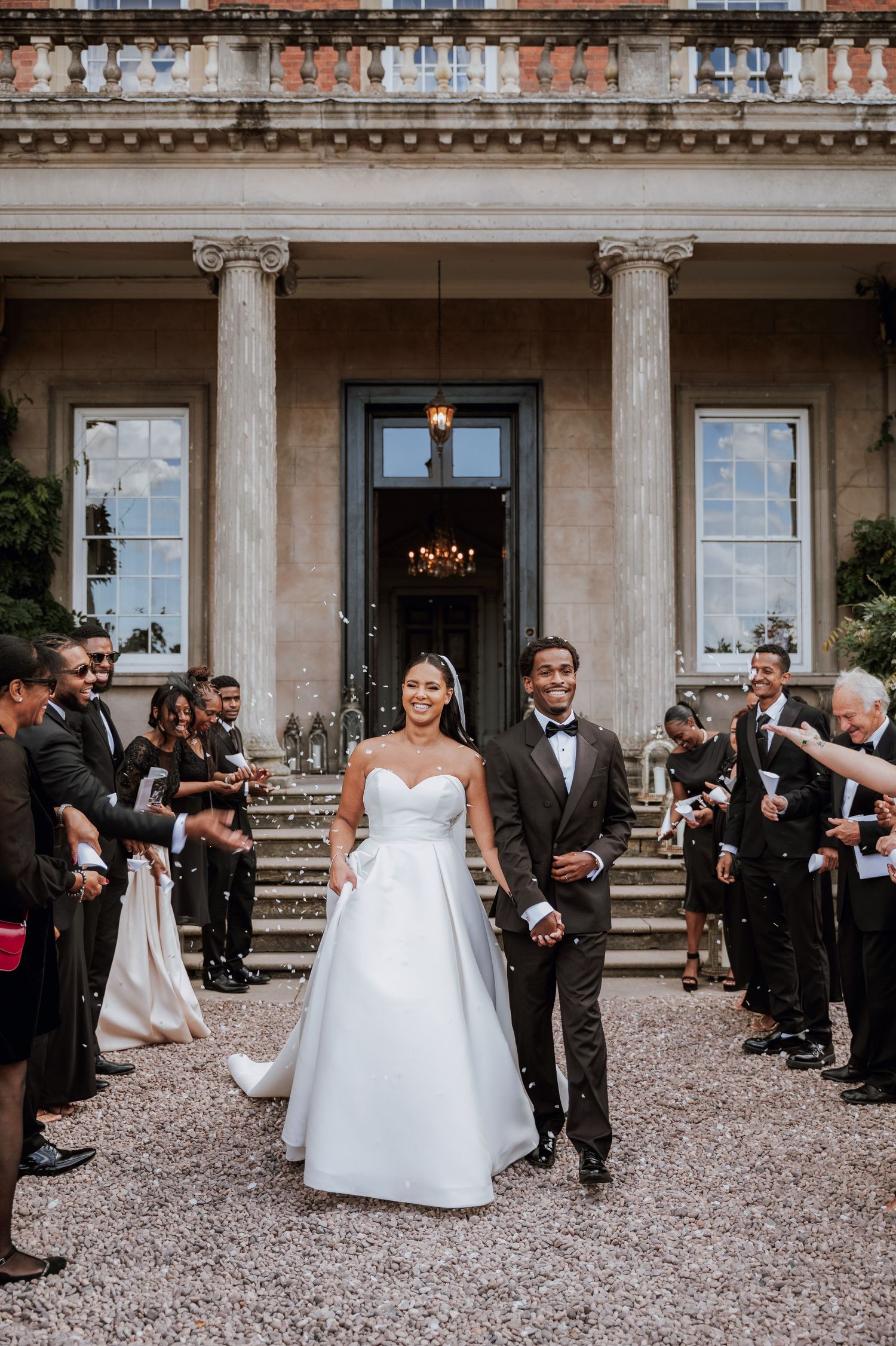 Wedding party in formal attire gathers outside a grand historic building with columns and gravel walkway.