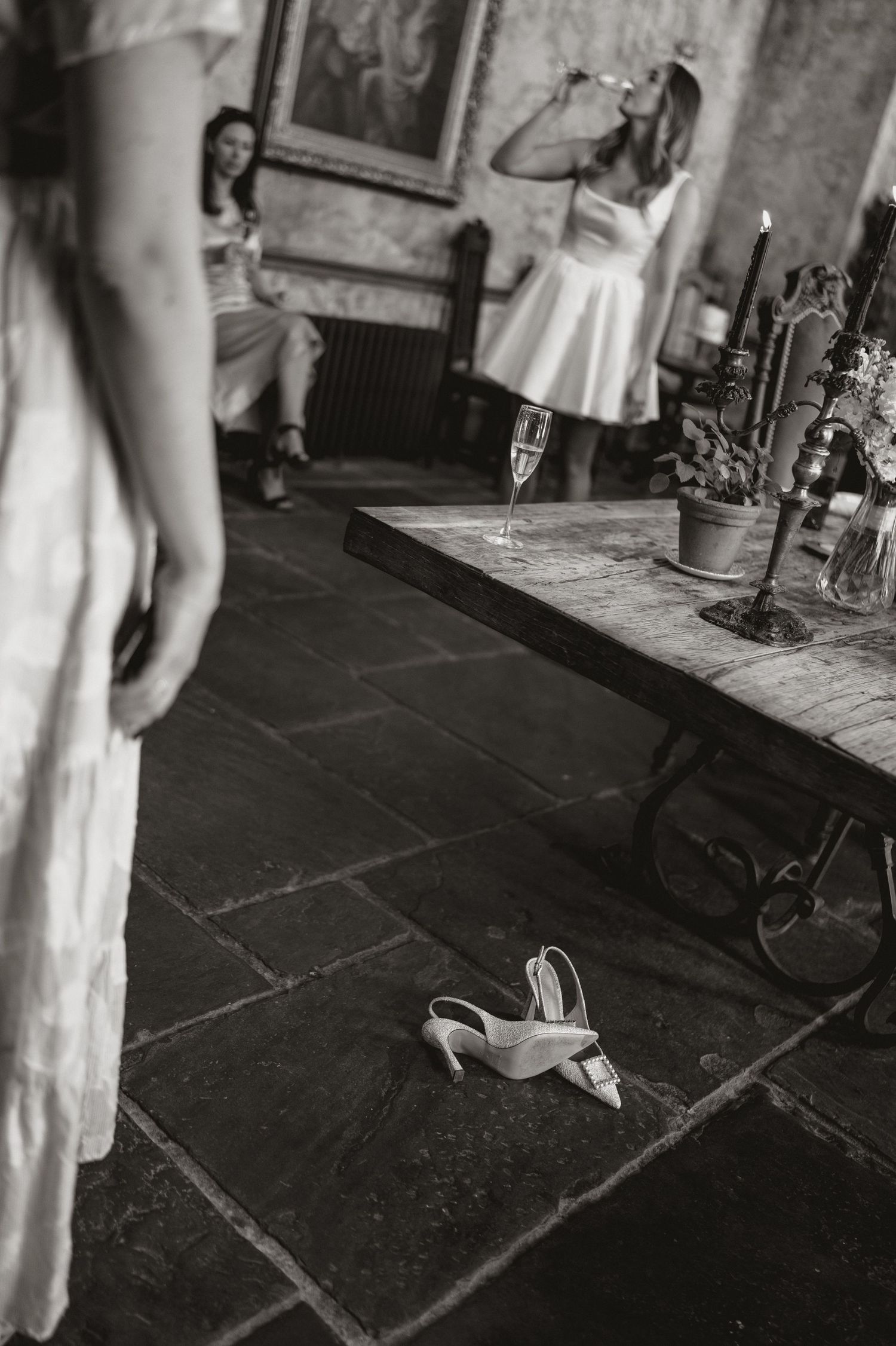 Black and white photo showing discarded high heels on stone floor with people in white dresses in background.