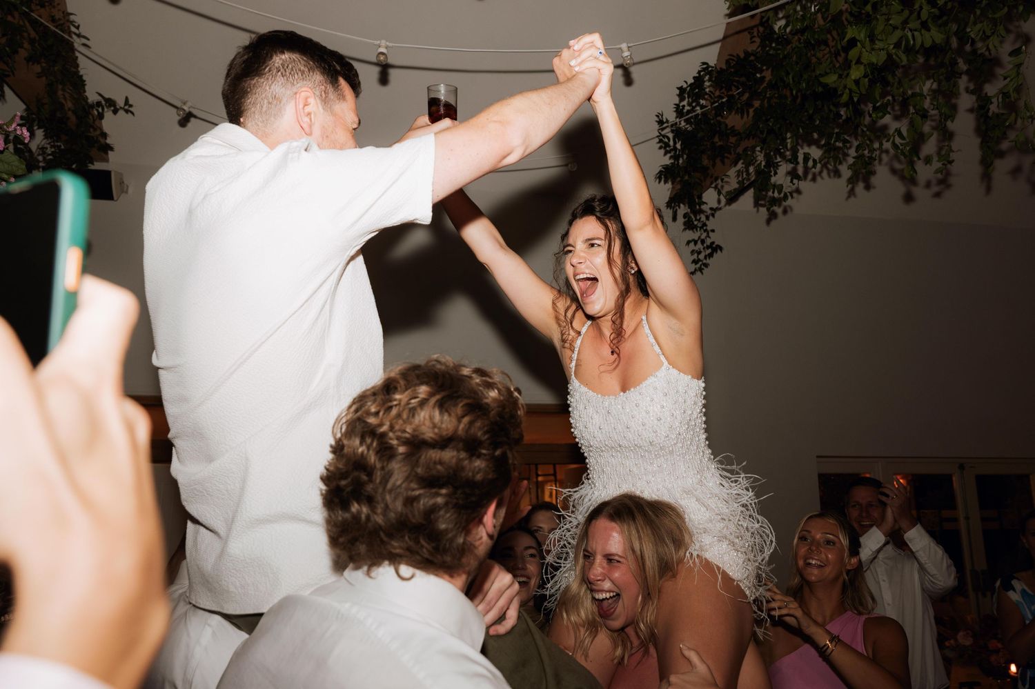 Energetic dance moves on a crowded wedding reception dance floor as guests cheer and take photos.