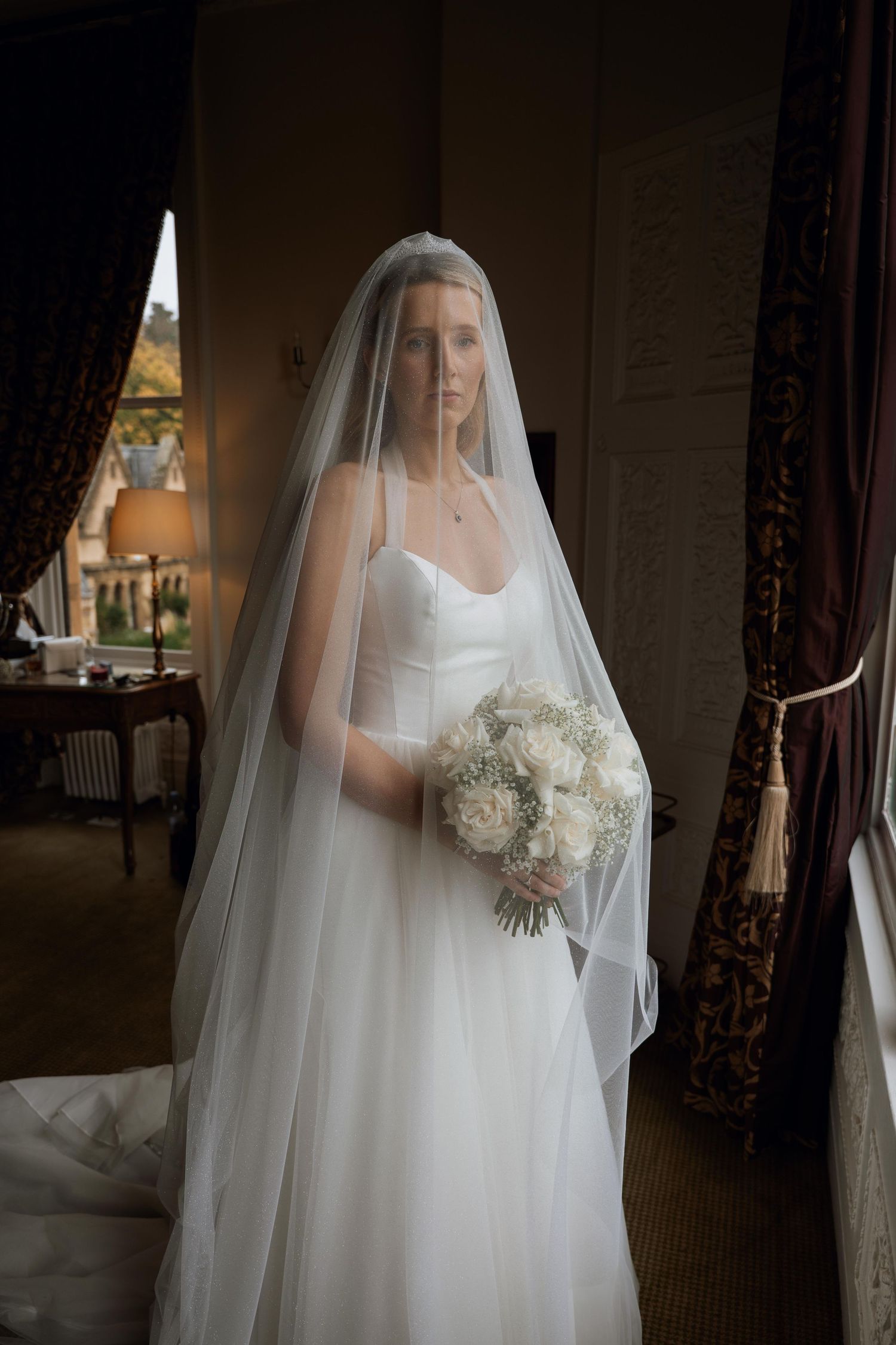 A bride in a classic white wedding gown and flowing veil stands in elegant indoor lighting creating a romantic silhouette.