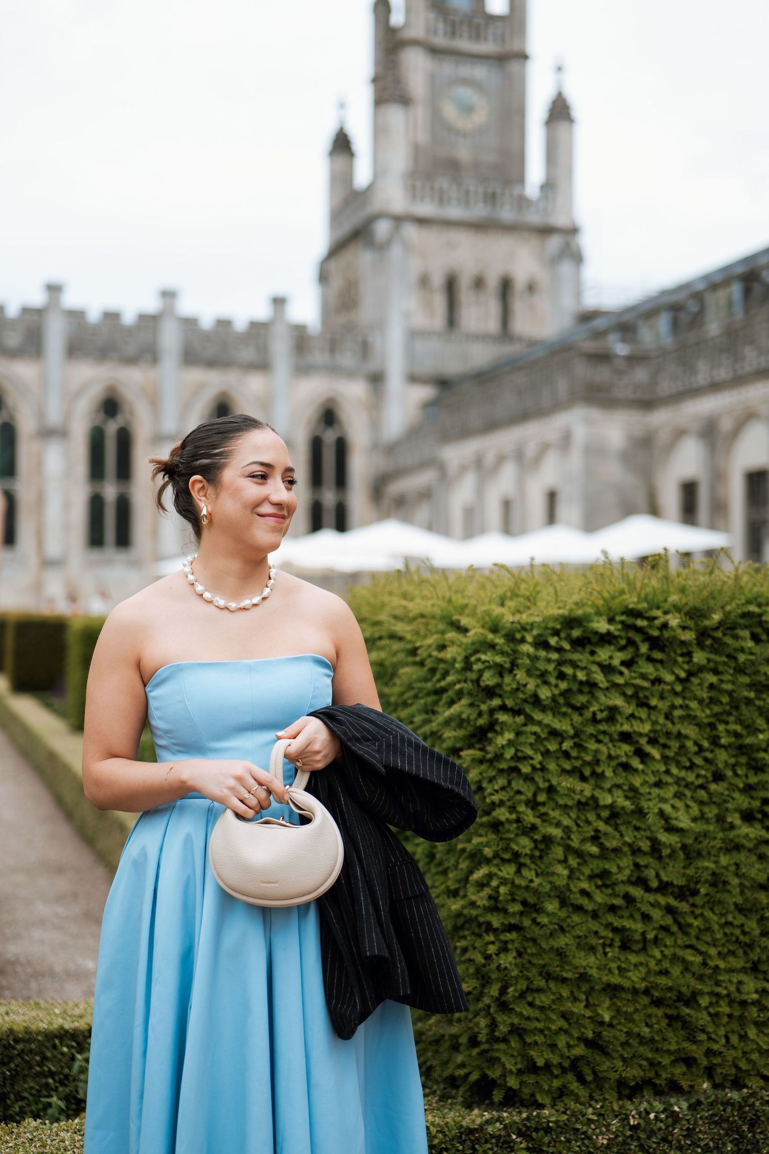 A person in a light blue strapless dress stands smiling near gothic architecture and manicured hedges.