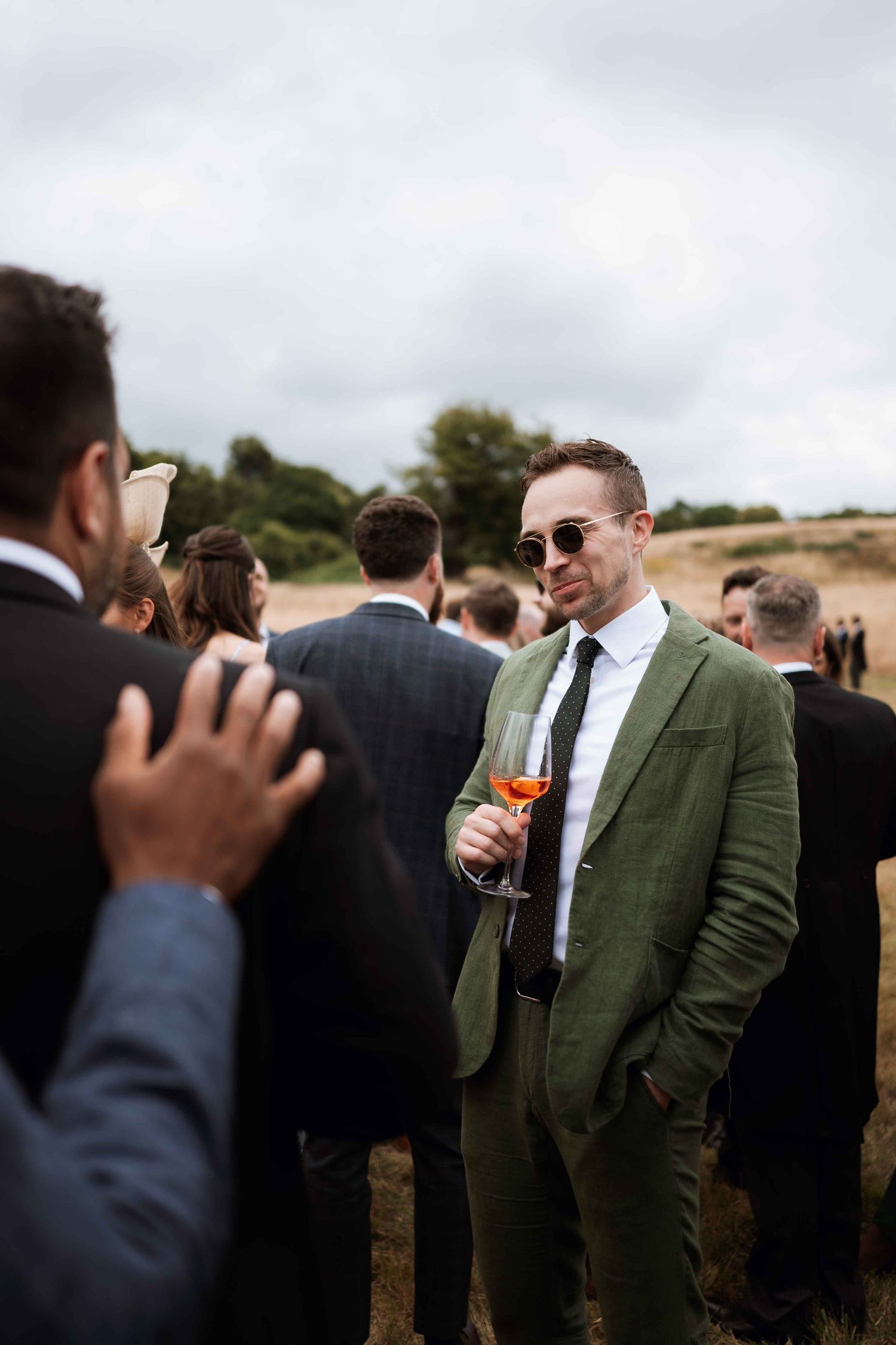 Wedding guests socialize outdoors with someone in a stylish olive green suit holding a cocktail.