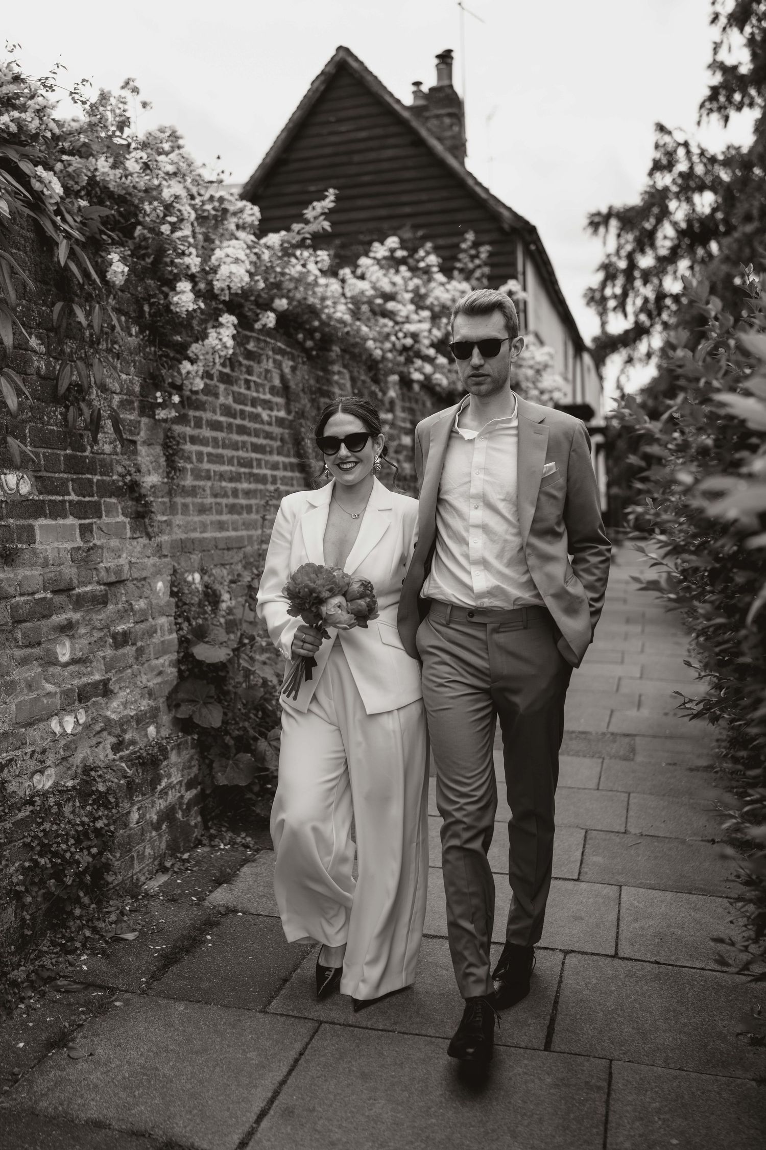 Black and white photo of a stylish couple walking together on a garden path wearing elegant suits and sunglasses.