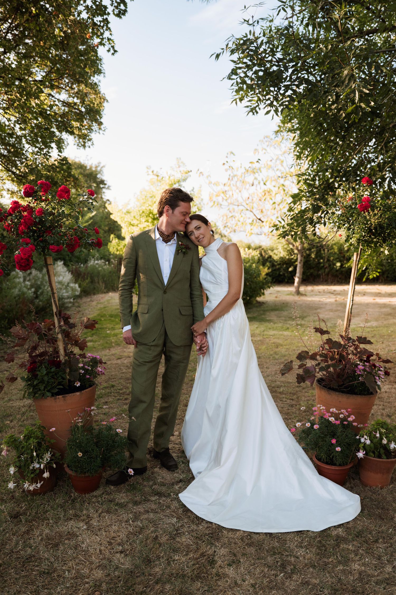 A couple in wedding attire poses together in a garden setting with red flowers and trees at sunset.