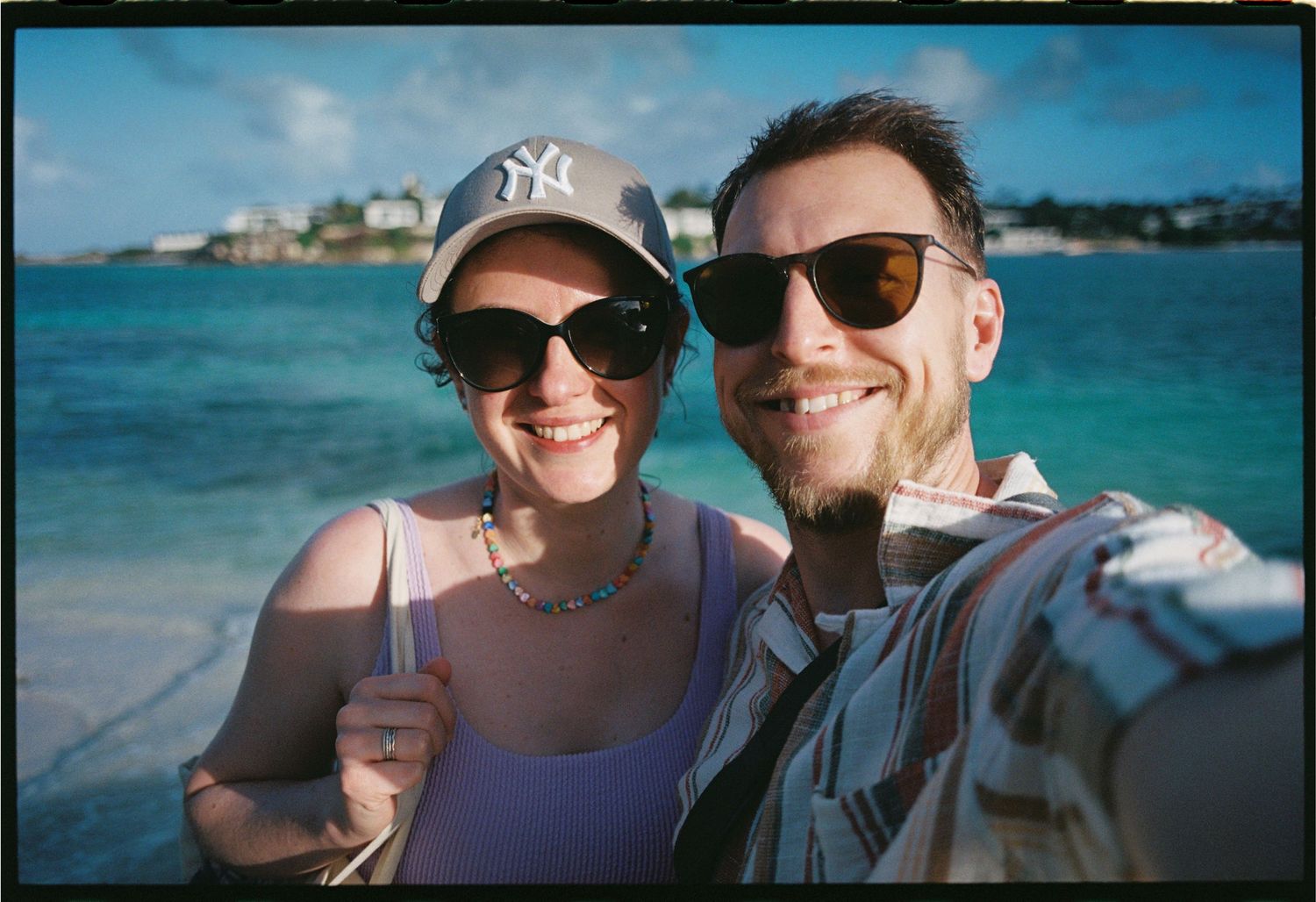 Smiling couple taking a selfie on a sunny beach with turquoise water, woman wearing NY cap and sunglasses.
