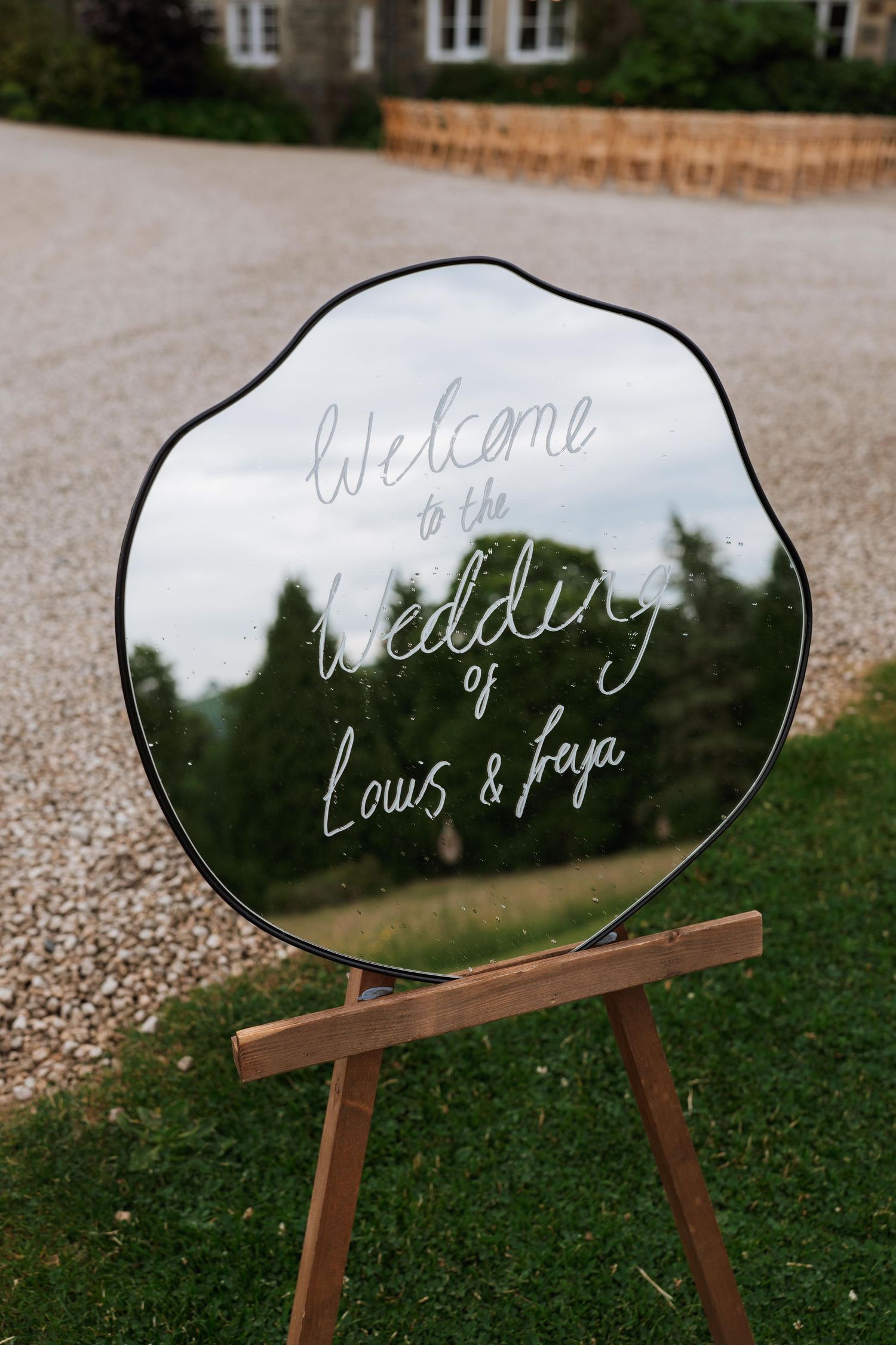 Mirrored welcome sign on wooden easel displays elegant white handwritten text for wedding decor outdoors.
