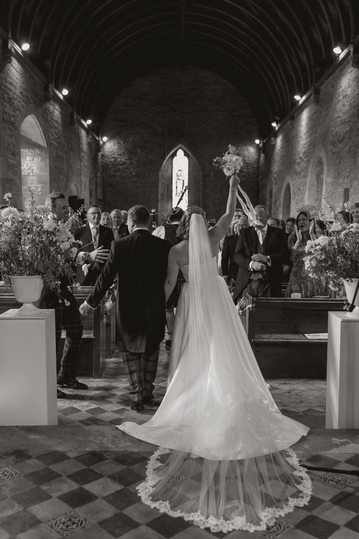 A black and white wedding ceremony in a stone church with a long veil trailing behind the bride down the checkered aisle.