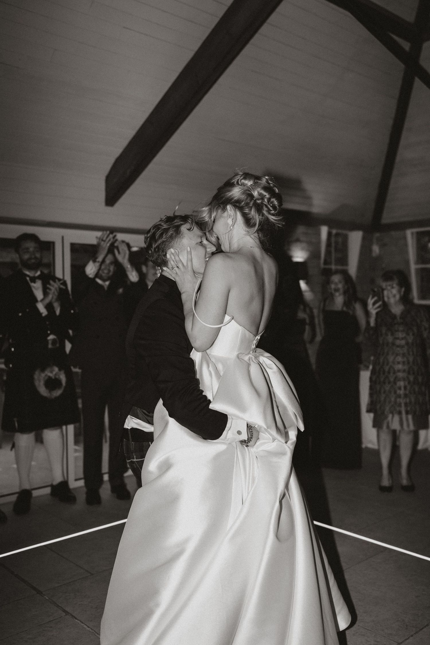 A black and white photograph captures a couple dancing intimately at a wedding reception with onlookers in the background.