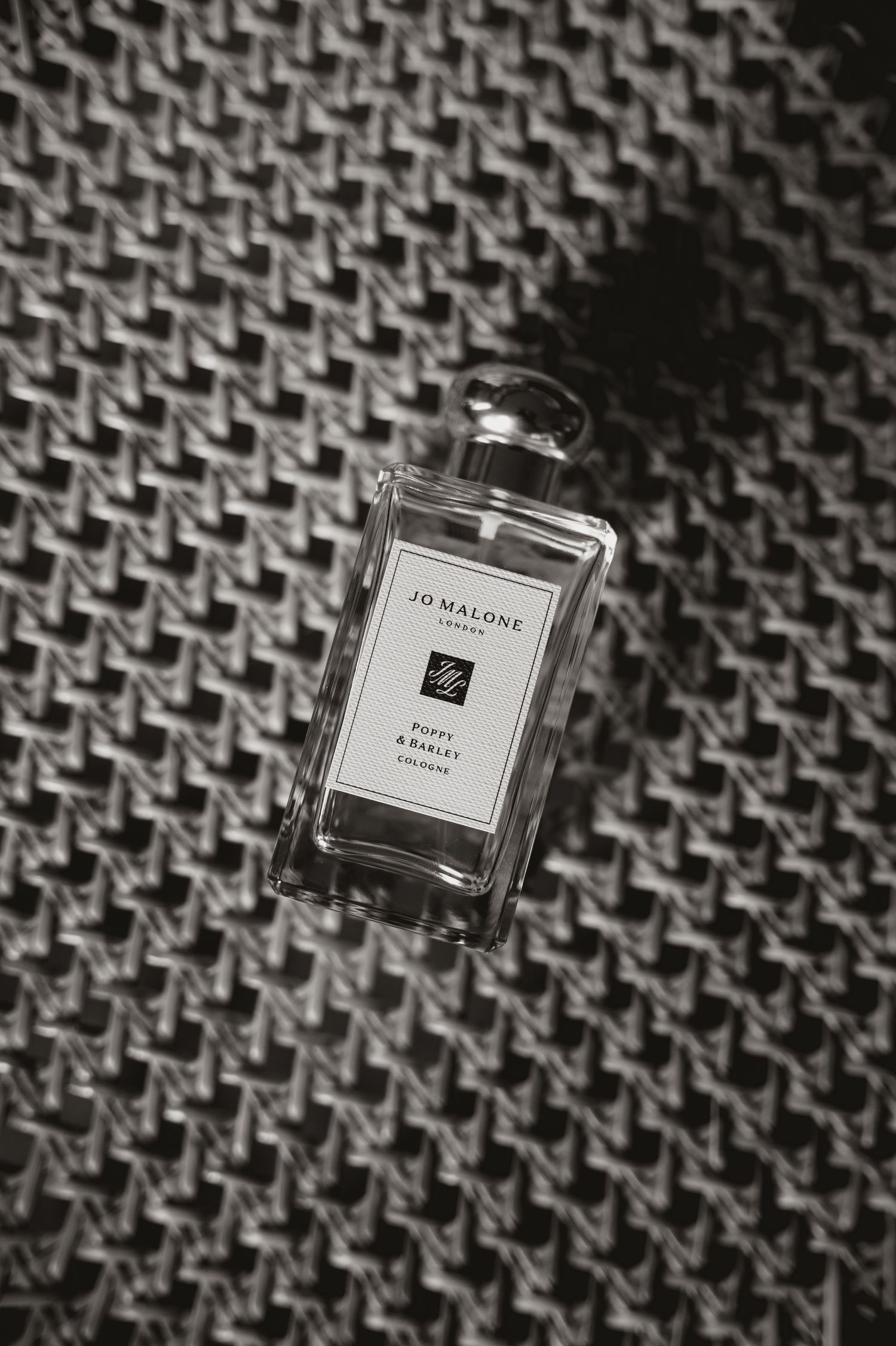 A black and white photograph of Jo Malone perfume bottle on textured fabric with diagonal pattern.