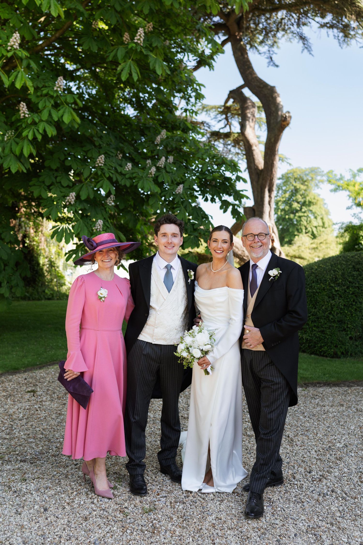 A group in formal wedding attire poses outdoors on a gravel path with trees and greenery in the background.