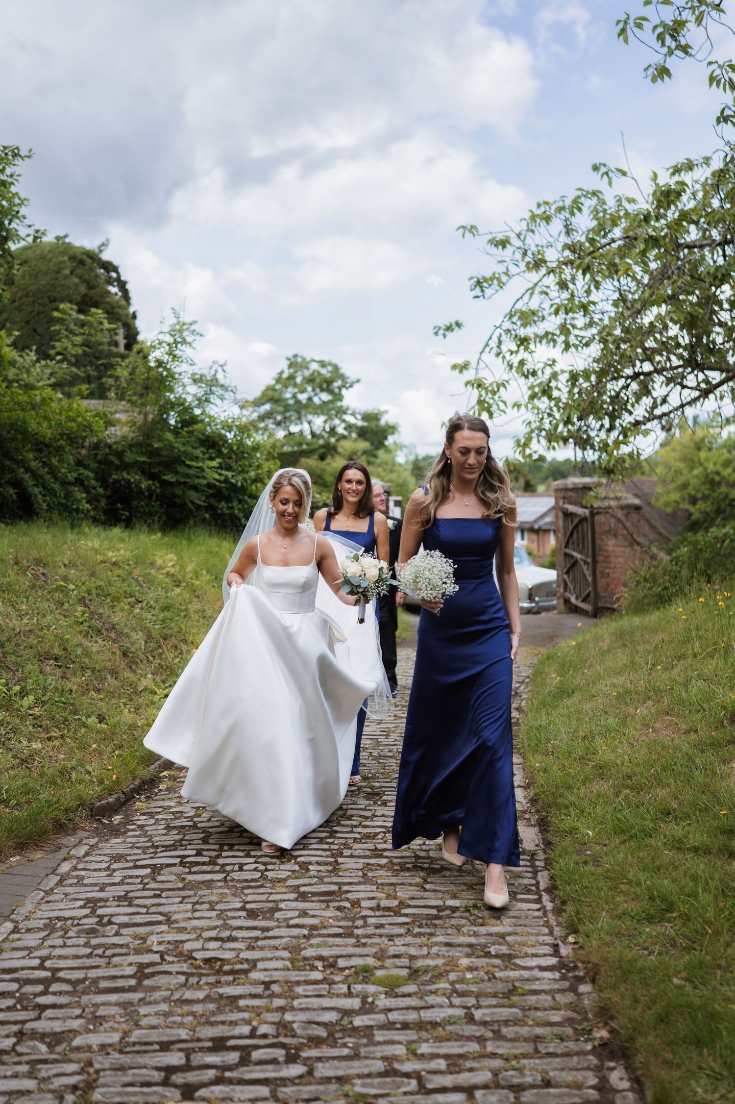 Wedding party walks up a cobblestone path surrounded by greenery on a cloudy day.