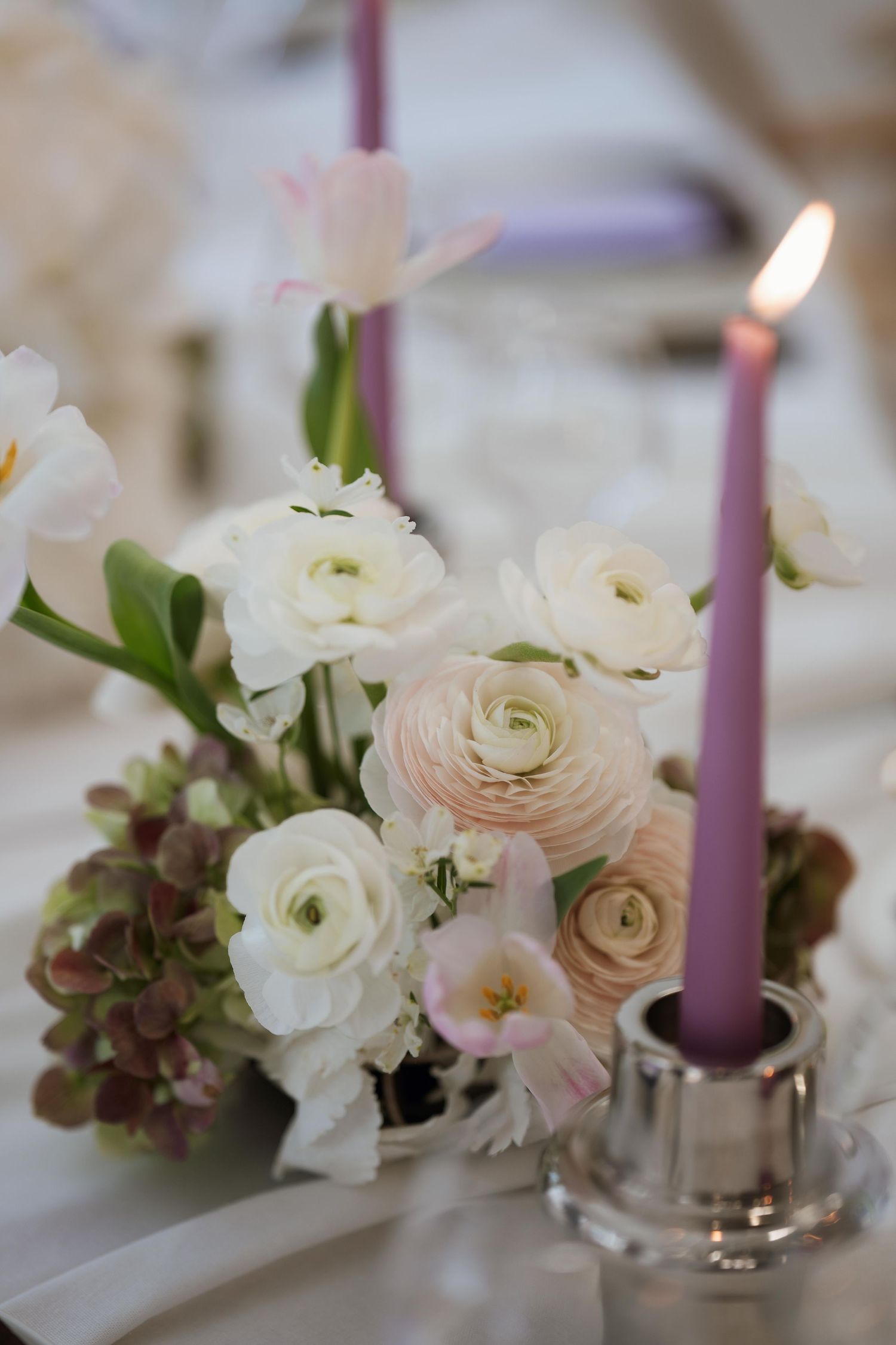 An elegant wedding centerpiece arrangement with white and blush ranunculus flowers and purple taper candles on a table setting.