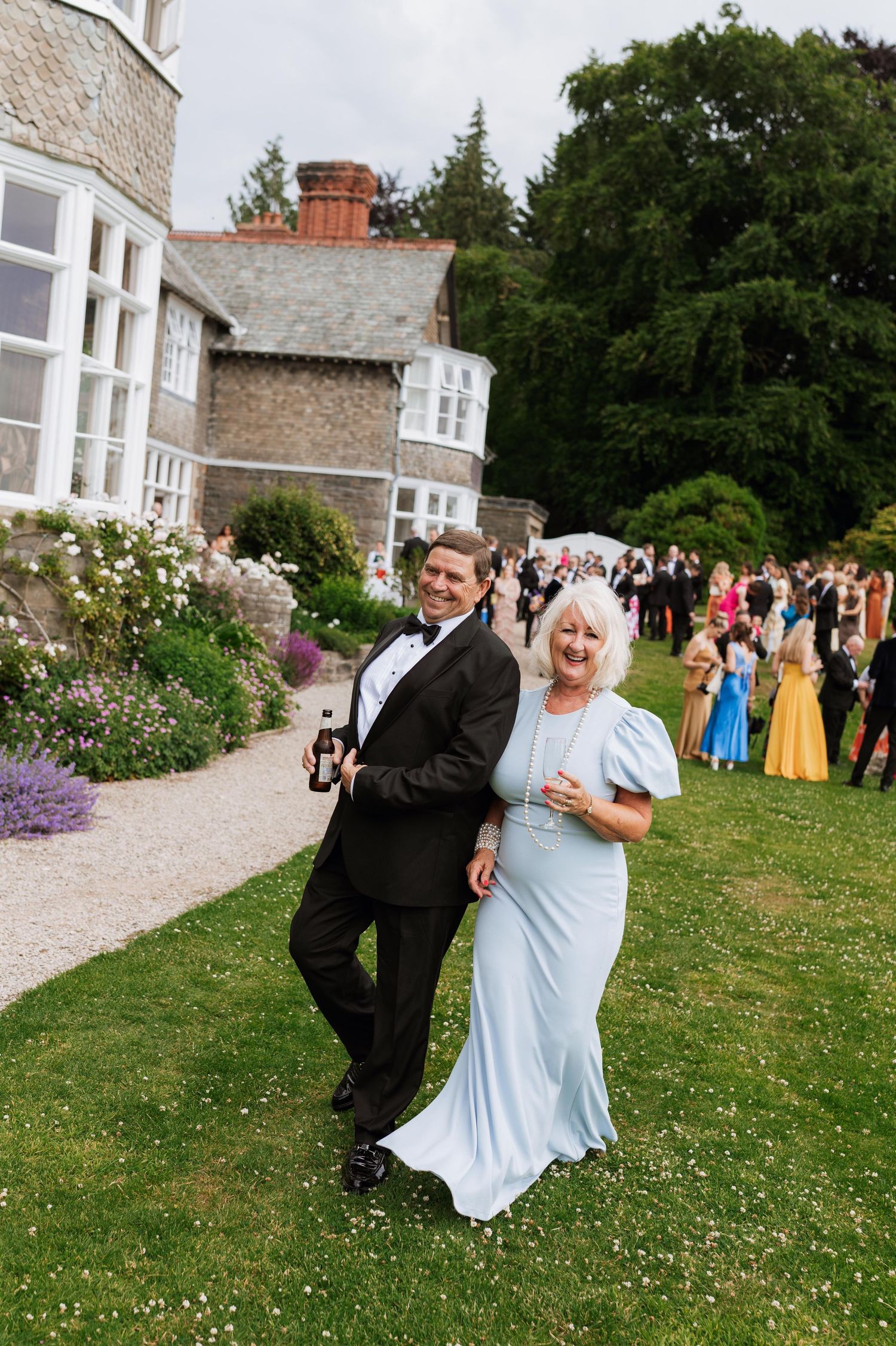 A couple in formal attire dances playfully on a grassy lawn outside a stone building during a wedding celebration.