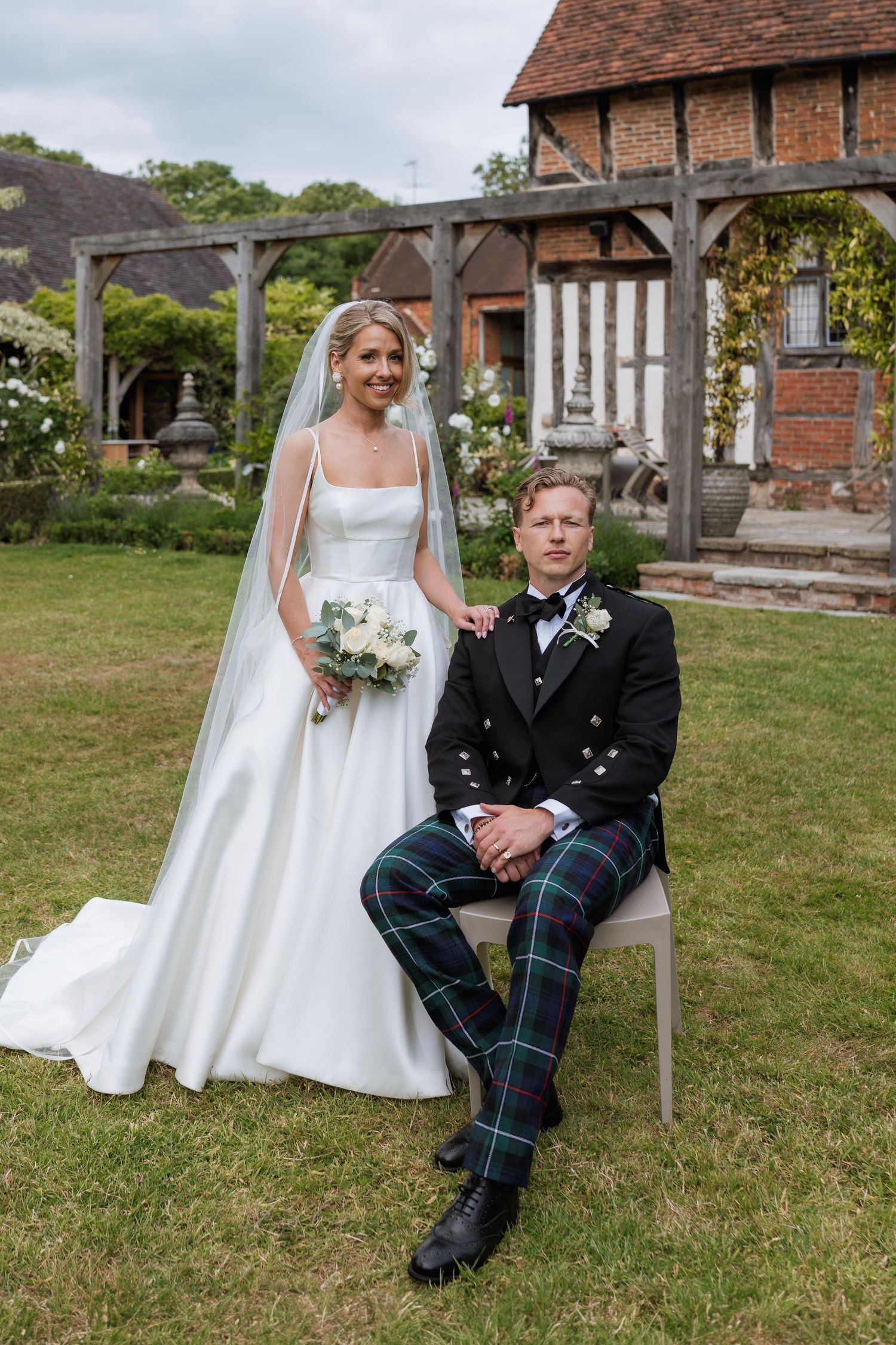 A couple in wedding attire sits on a bench outdoors, with the groom wearing a traditional Scottish kilt.