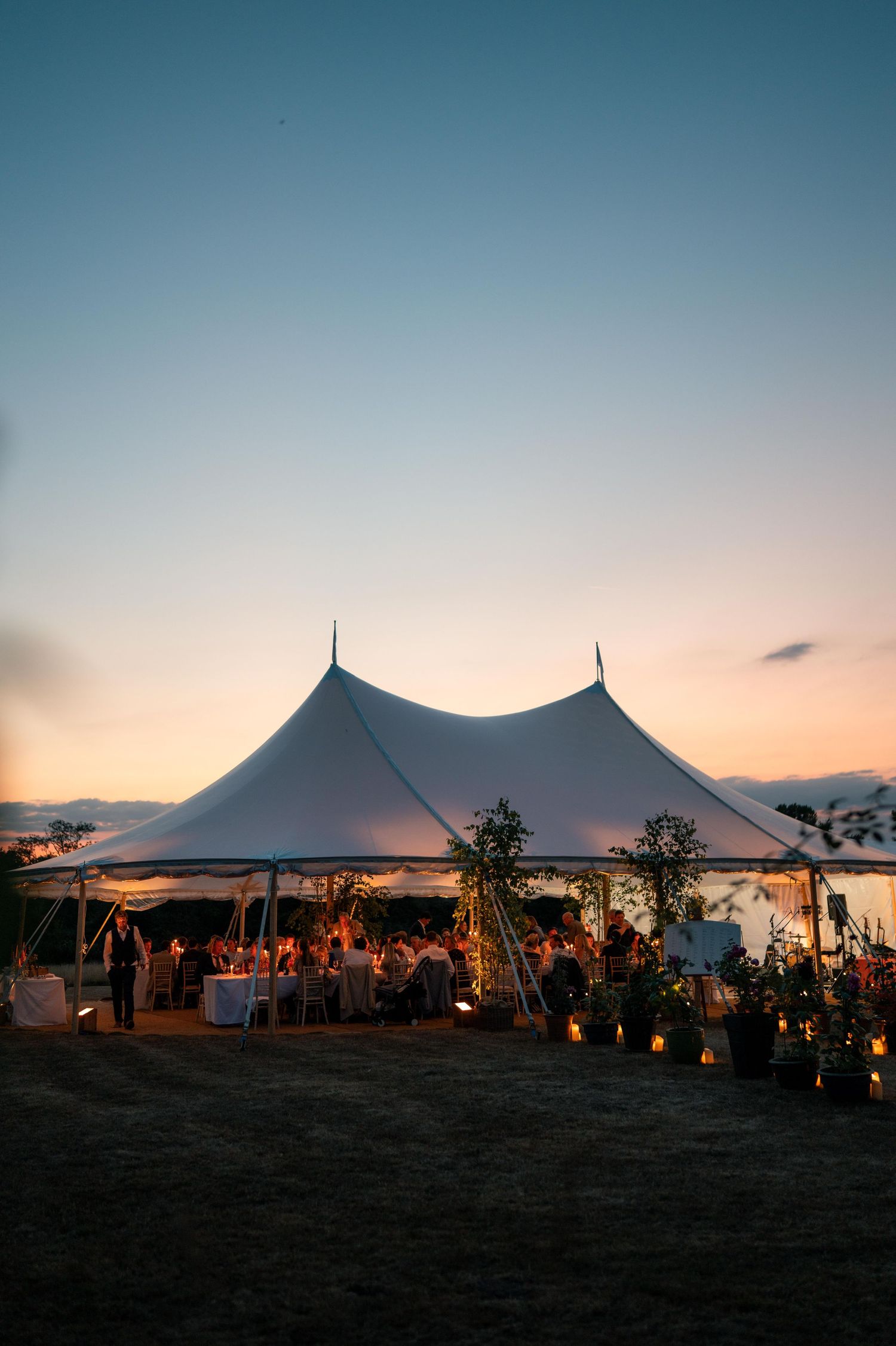 White wedding tent silhouetted against a sunset sky during an outdoor reception with glowing lights and palm trees.