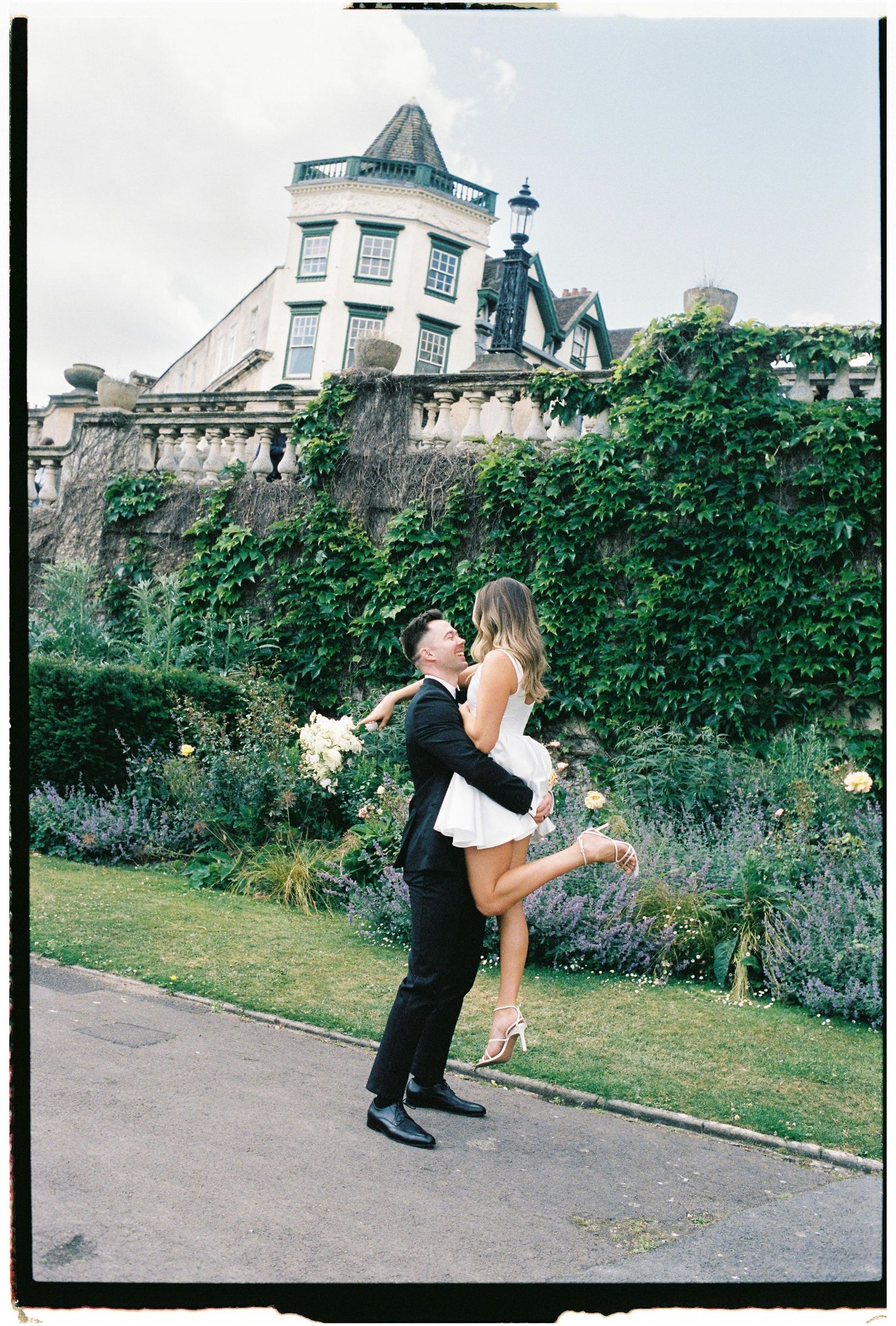 A couple embraces in a romantic moment on the lawn of an elegant Victorian mansion with ivy-covered walls.