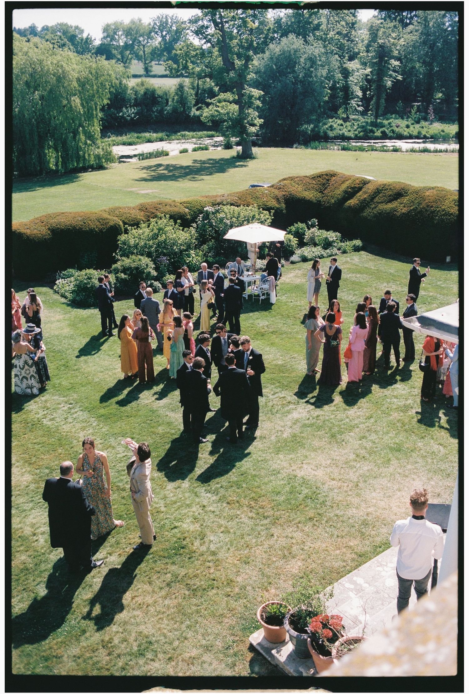 Outdoor garden party gathering on a sunny day with guests mingling on a grassy lawn near trees and a white tent.