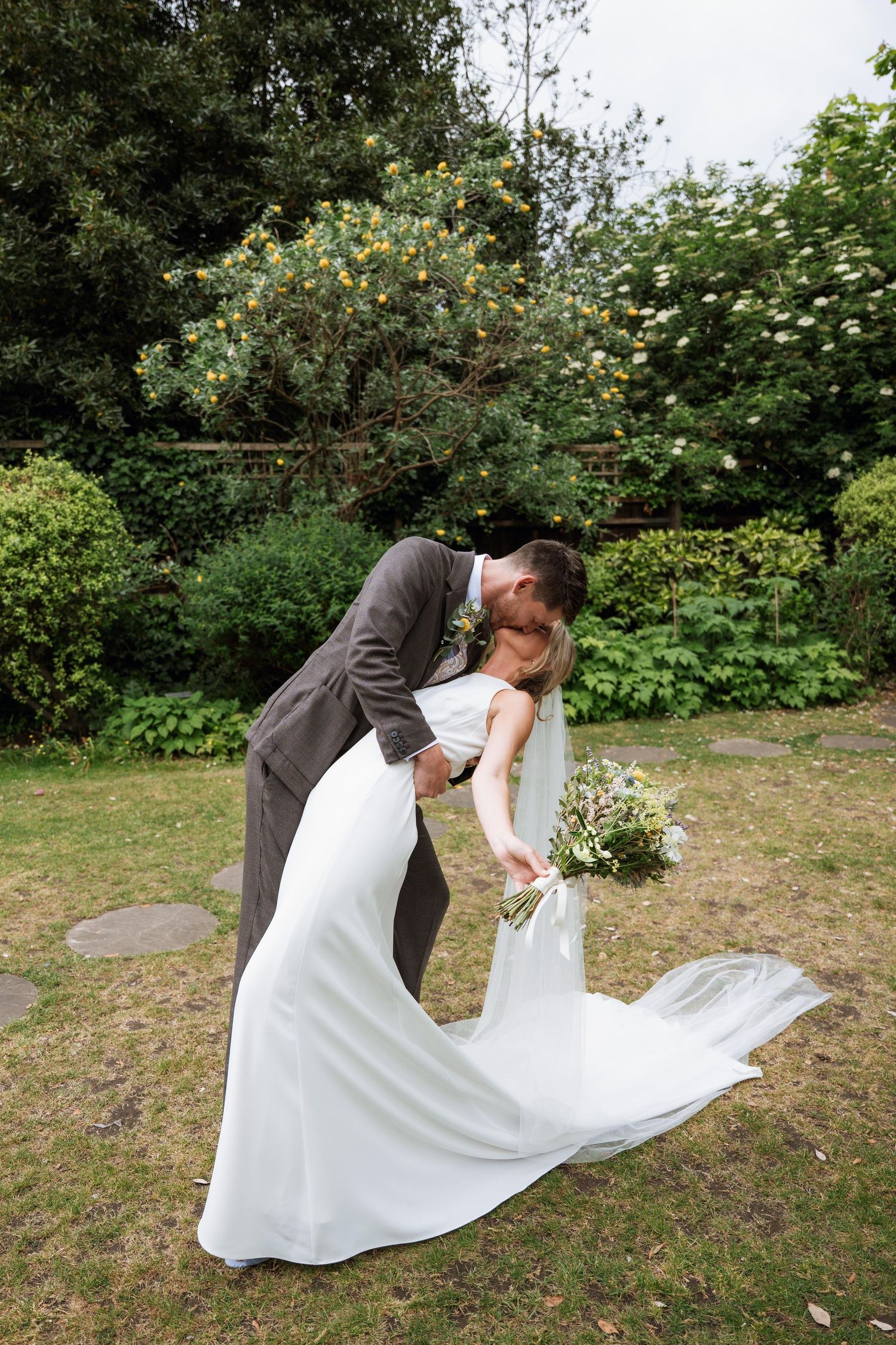 A couple in wedding attire share a romantic dip pose in a garden setting surrounded by greenery and flowers.