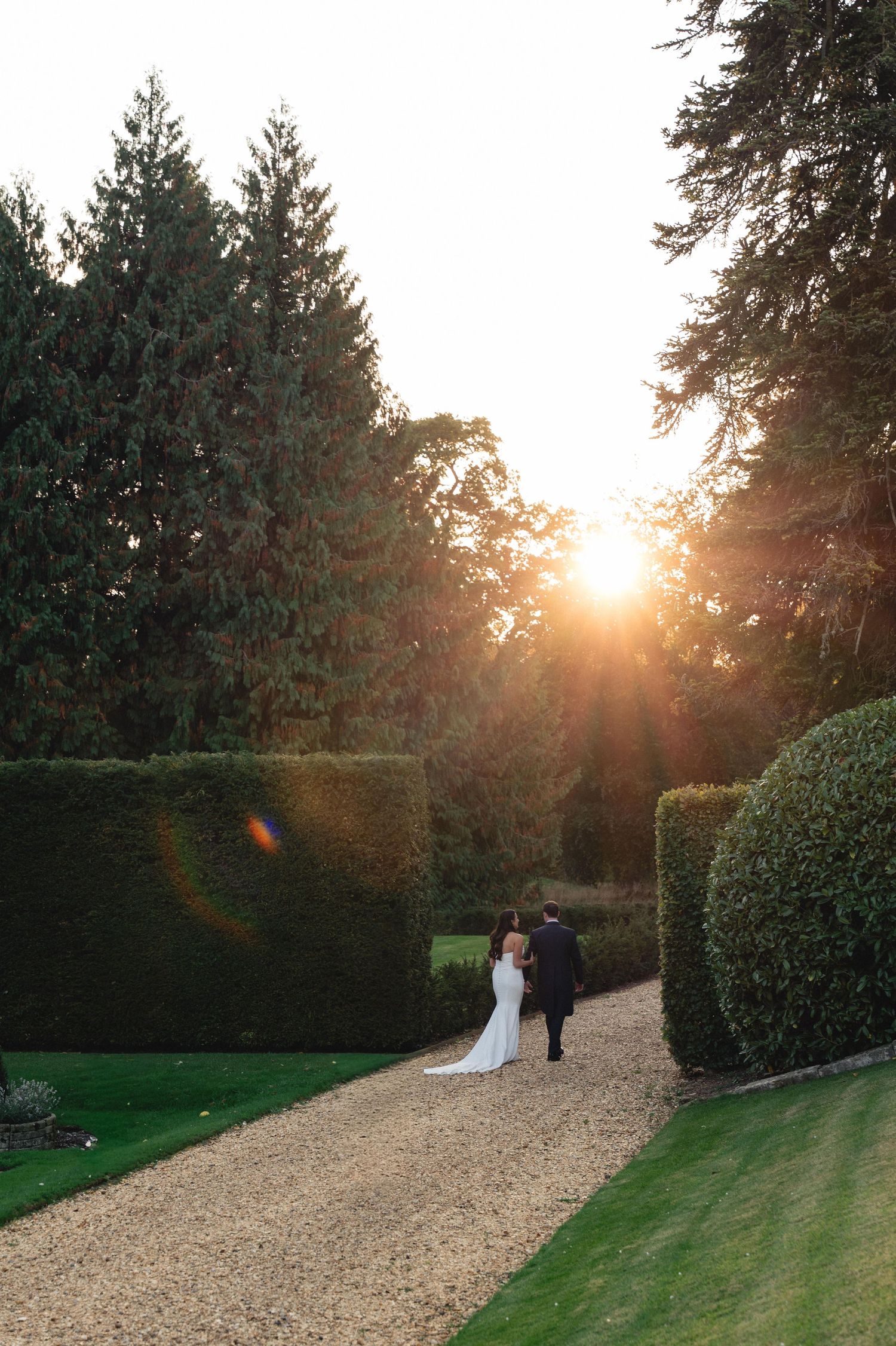 A couple walks along a gravel path in a garden at sunset with evergreen trees and manicured hedges in the background.