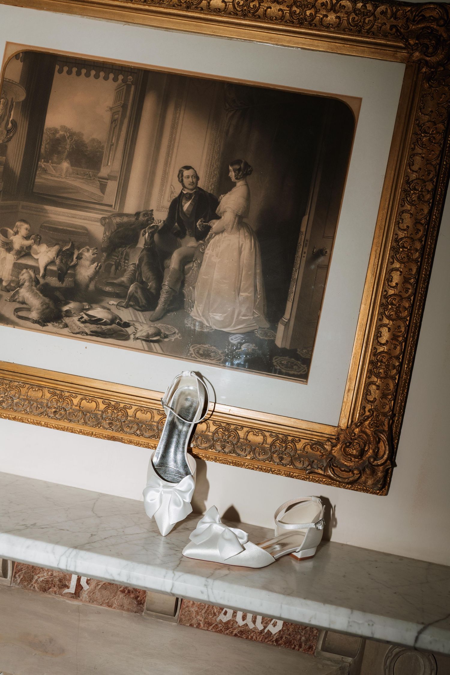 A series of ornate gold-framed artworks displayed on a white wall with elegant white heels placed in front of them.