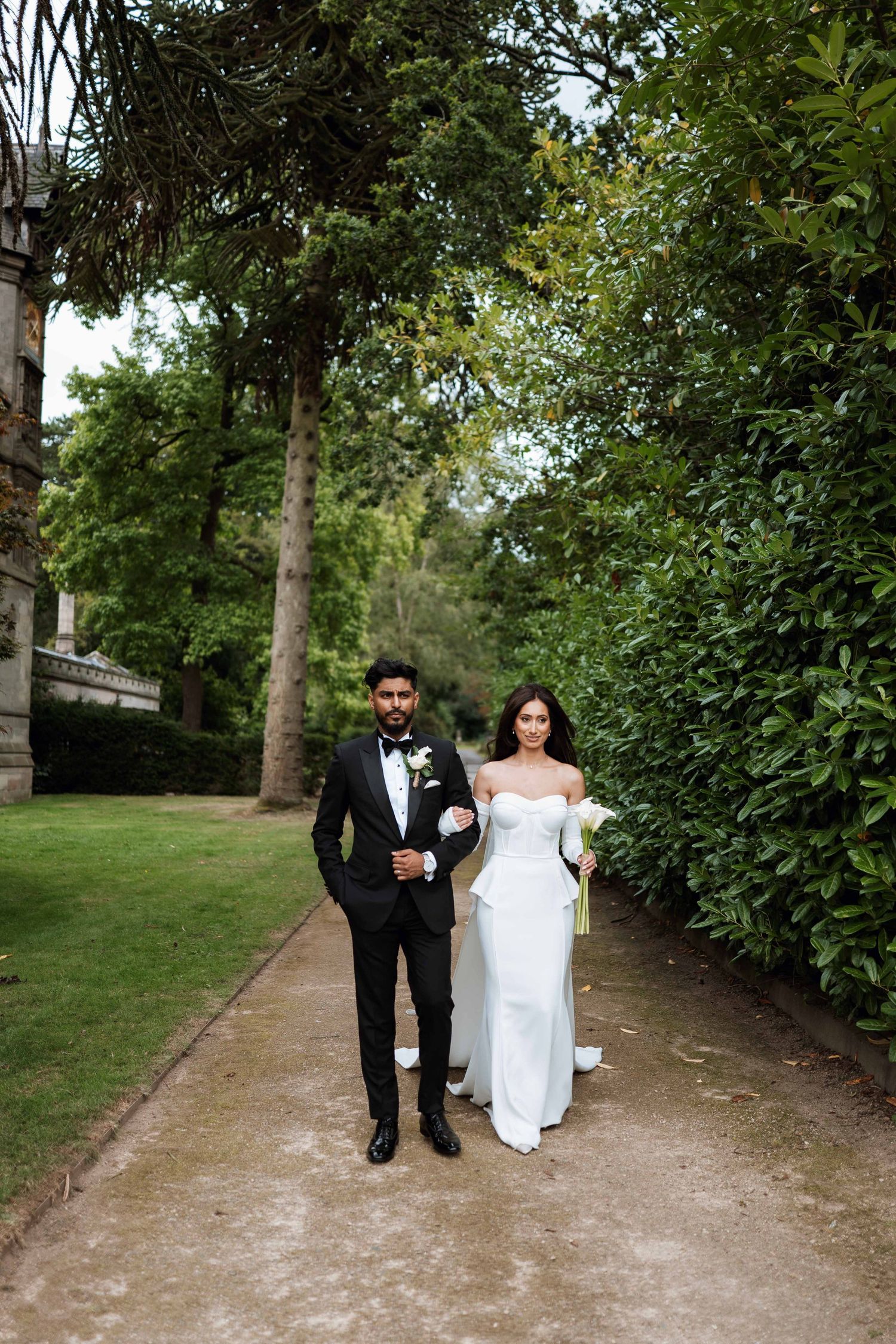 A couple in formal wedding attire walks along a tree-lined garden path on their special day.