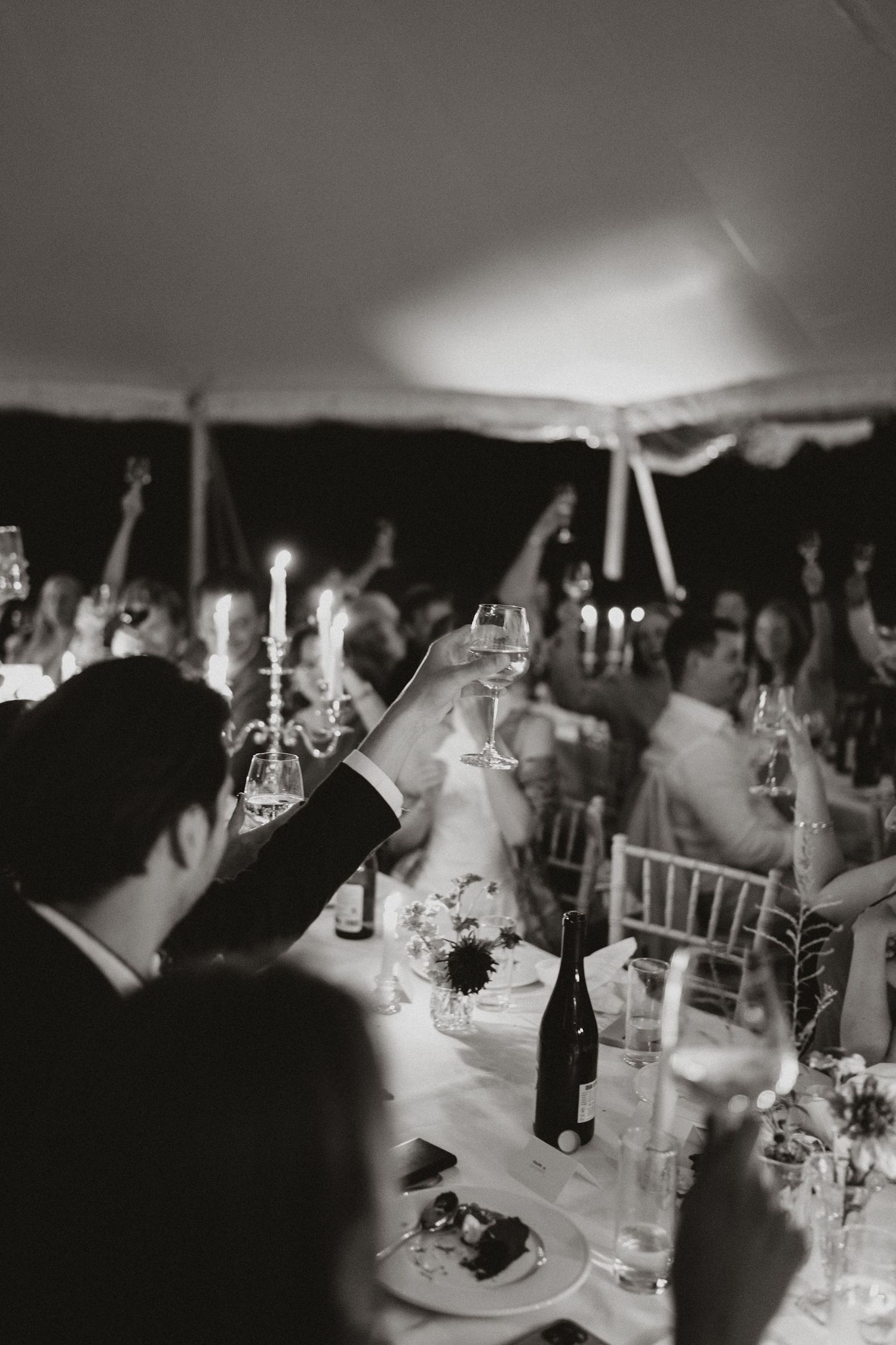 A black and white photo of guests raising wine glasses in a toast during an elegant wedding reception under a tent.