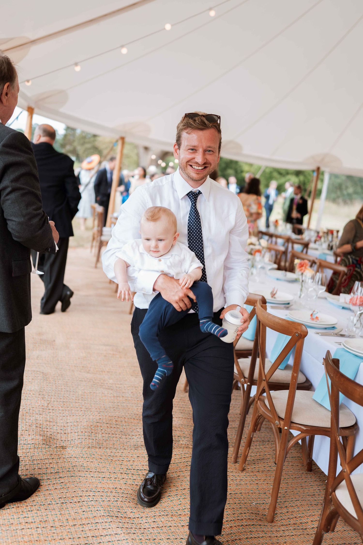 A person in formal attire holds a baby at an elegant tented wedding reception with table settings visible.