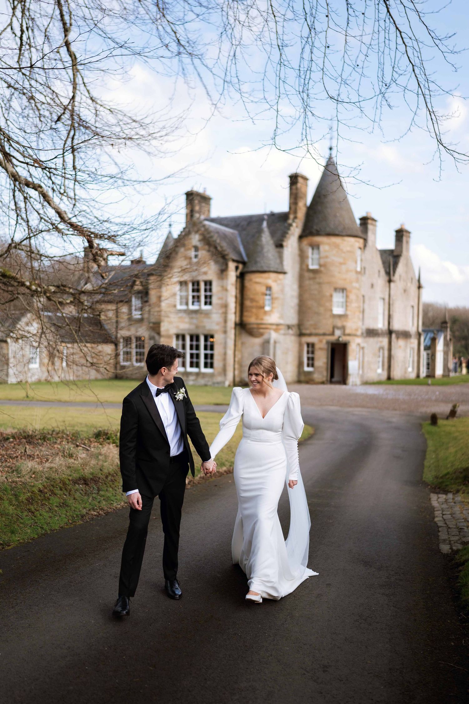 A couple walks along a driveway path leading to a beautiful Scottish castle-style estate on their wedding day.