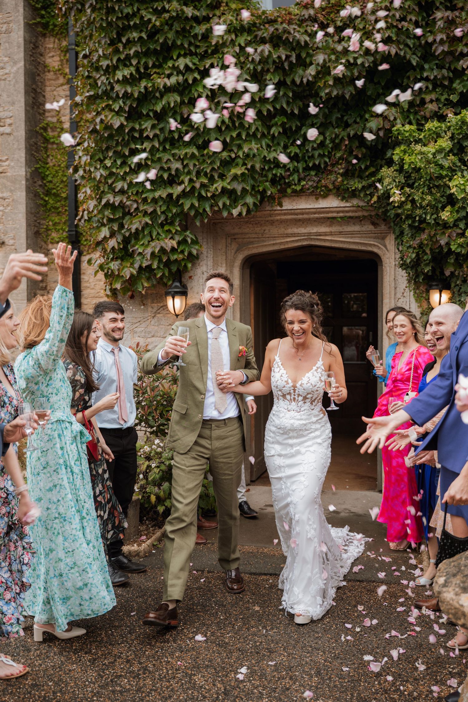 Wedding guests shower newlyweds with confetti outside a historic stone building covered in climbing vines and flowers.