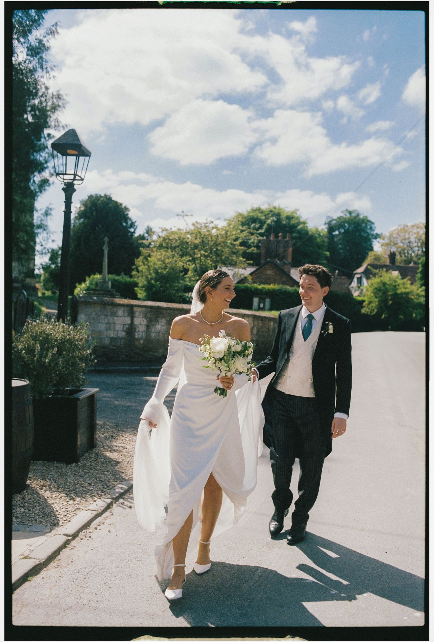 A newlywed couple walks along a sunlit path with a vintage lamppost and greenery in the background on their wedding day.