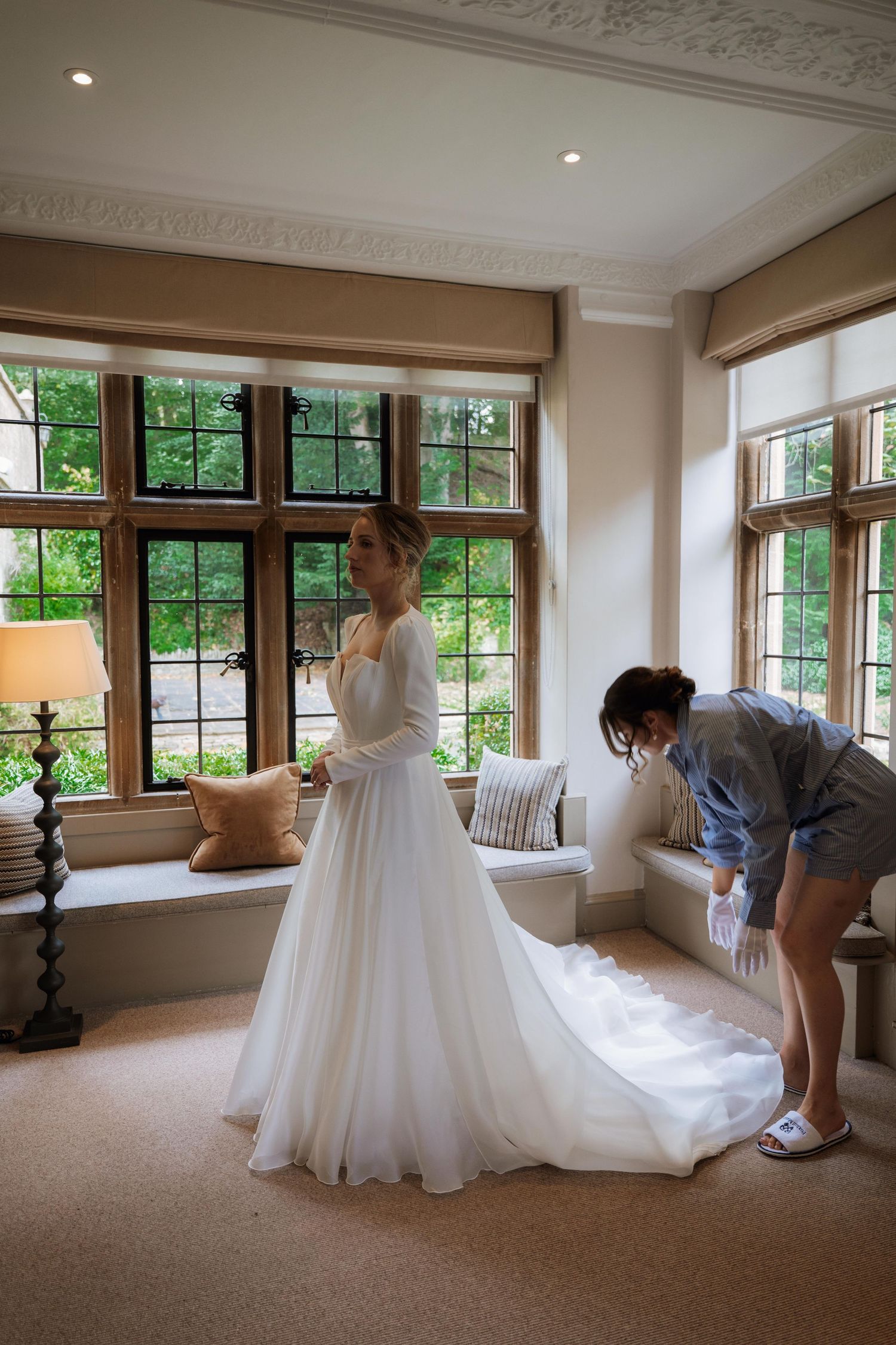 A wedding dress being prepared and adjusted in a bright room with large windows.