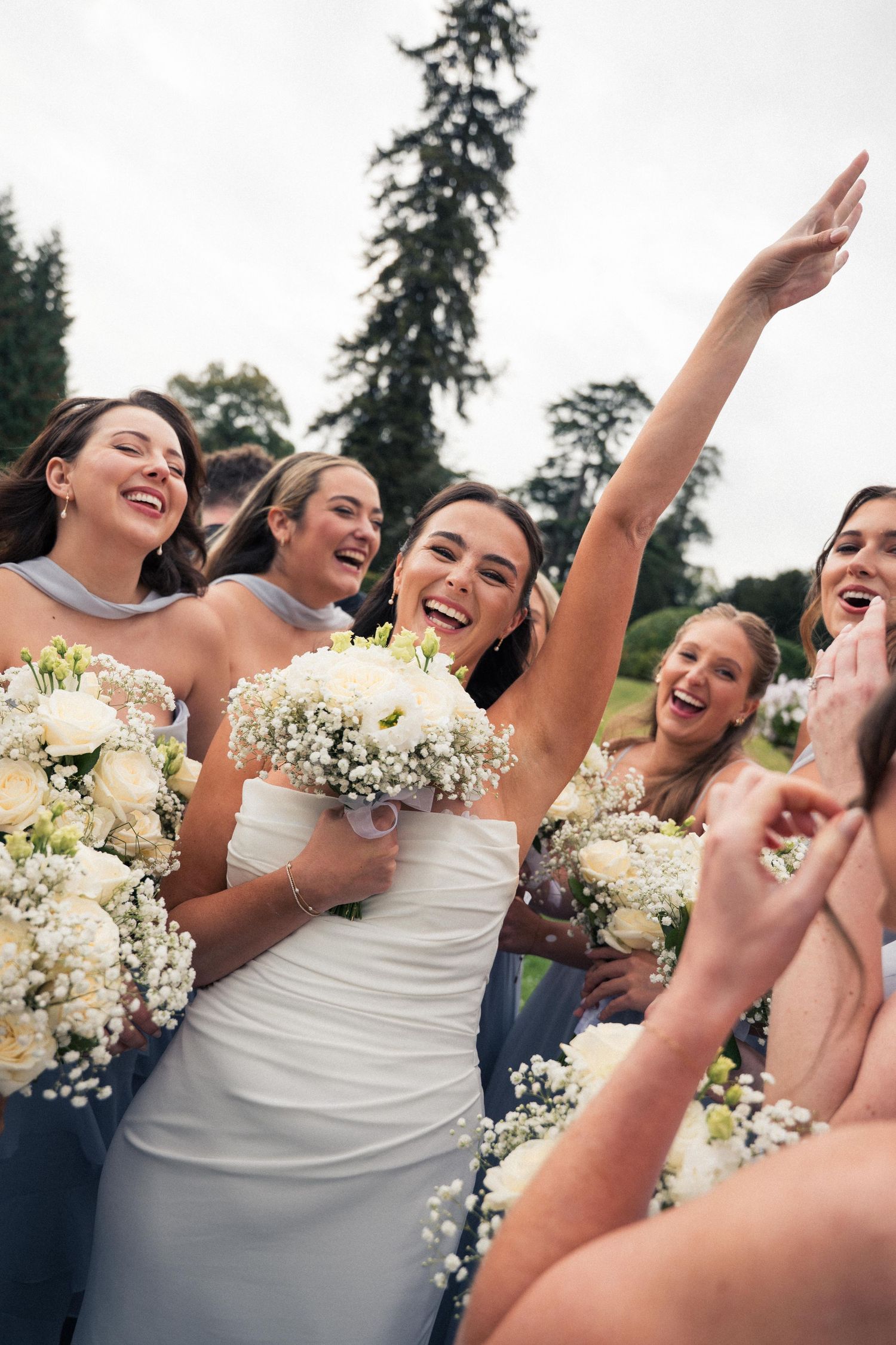 Joyful bridesmaids in blue dresses celebrate with white bouquets at an outdoor wedding celebration.