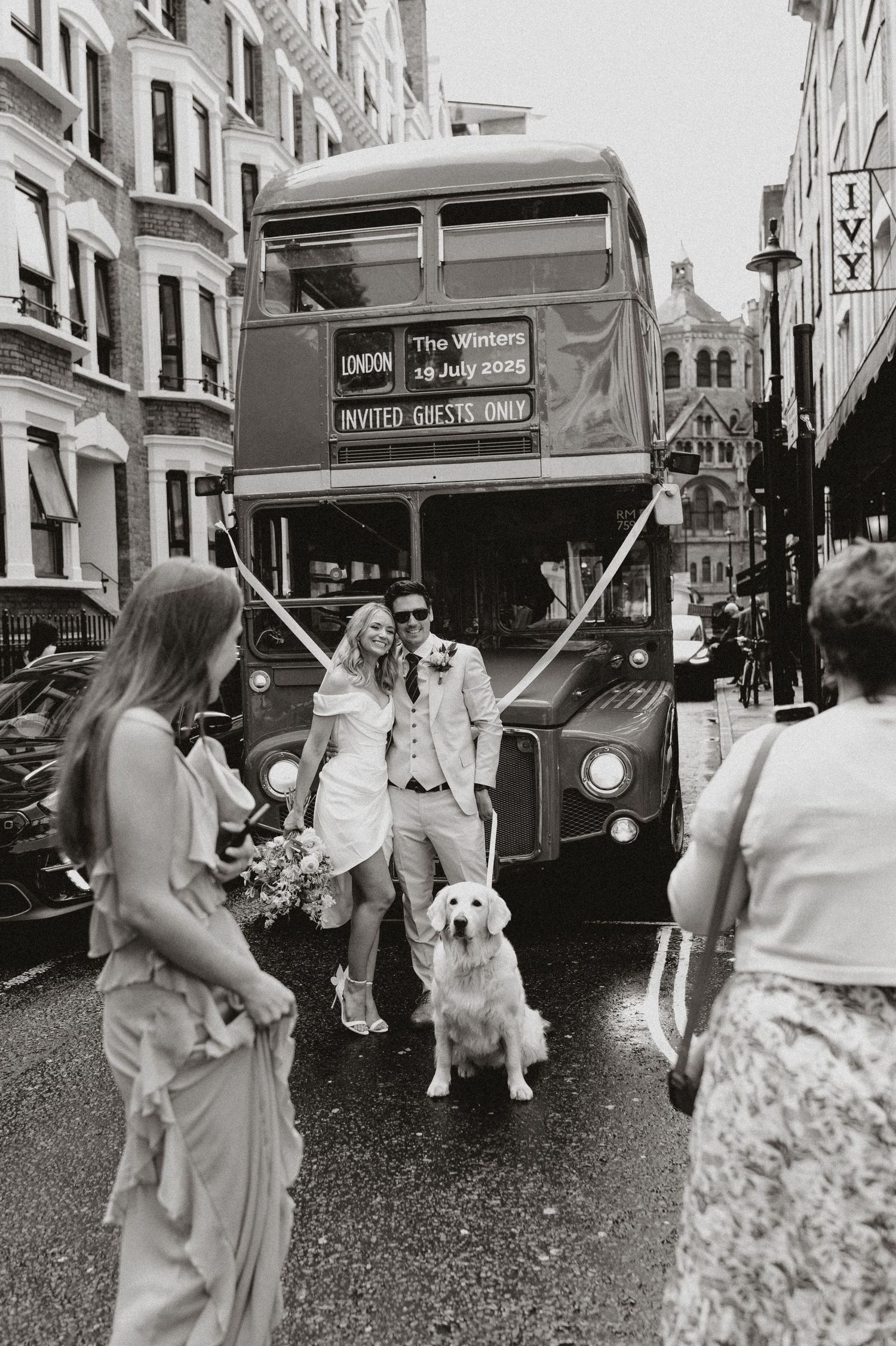 A vintage London double-decker bus with people and a golden retriever dog standing on a city street in black and white.