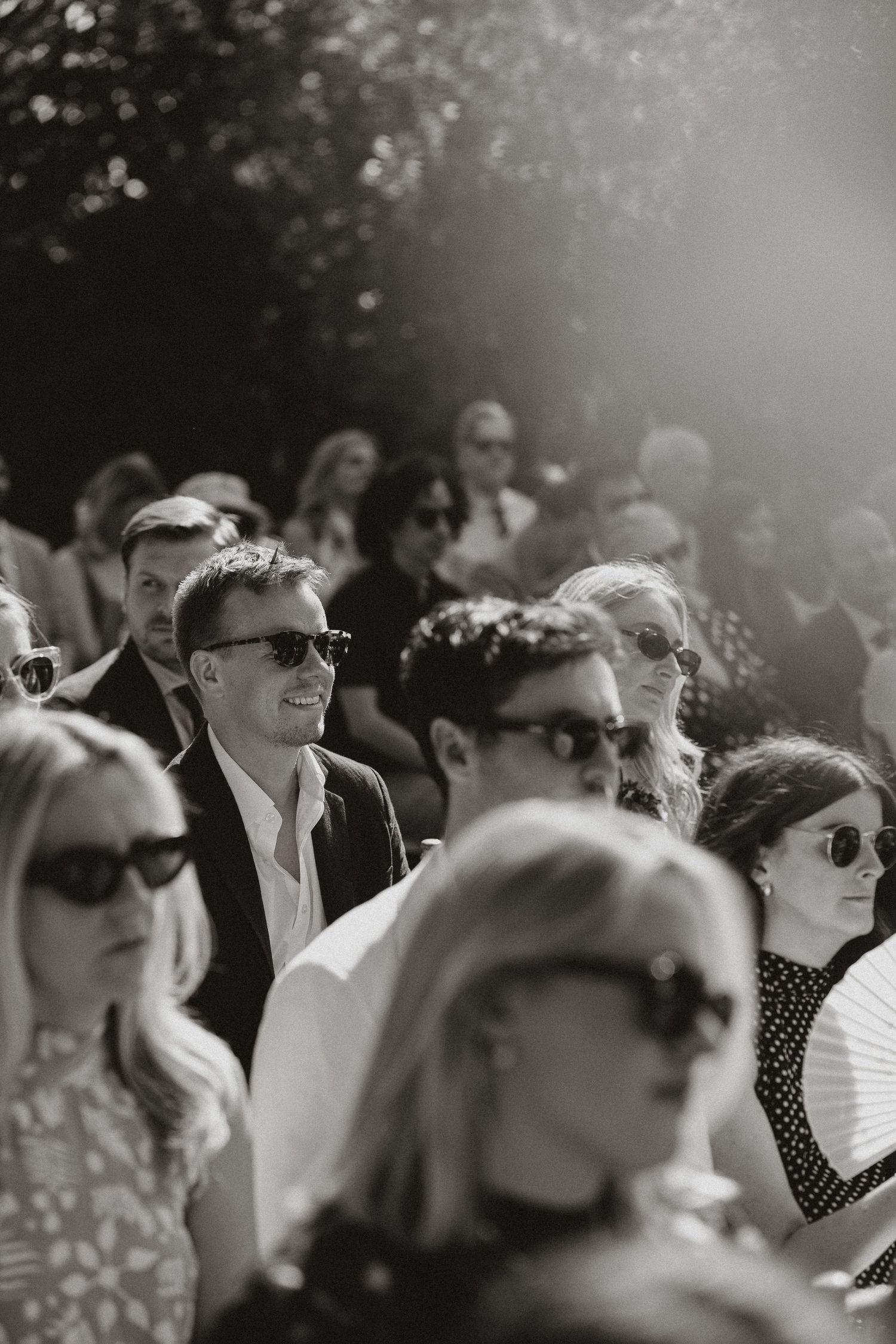 A black and white photo of wedding guests in sunglasses sitting outdoors on a sunny day.