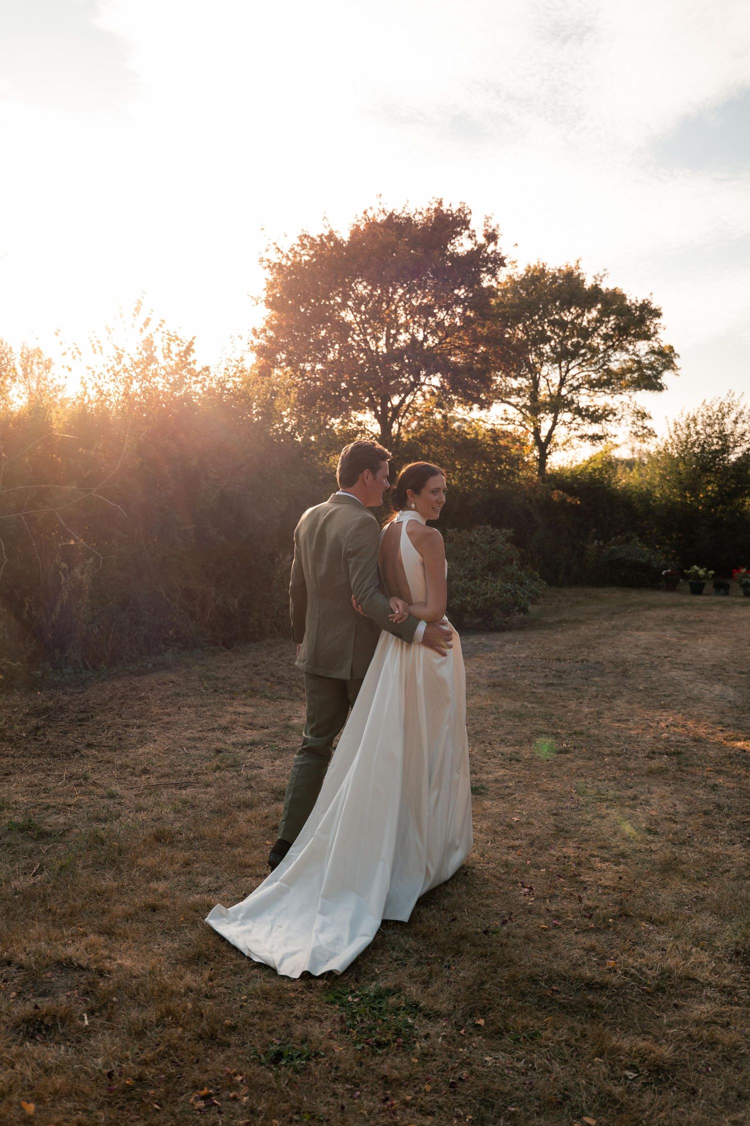 A couple in wedding attire embrace during golden hour sunset with silhouetted trees in the background.