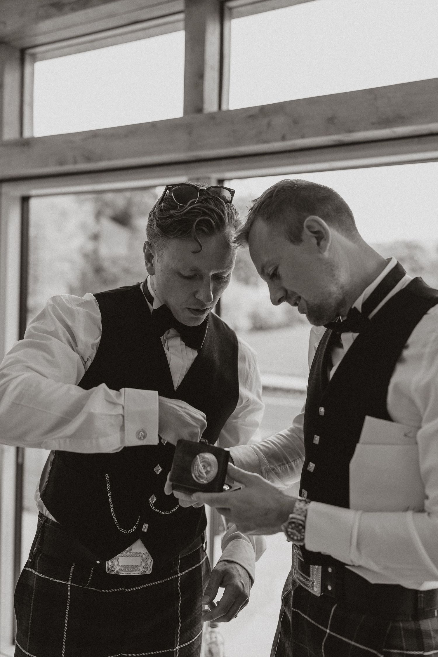 Two groomsmen in white shirts and black vests check their pocket watches together in a black and white photograph.