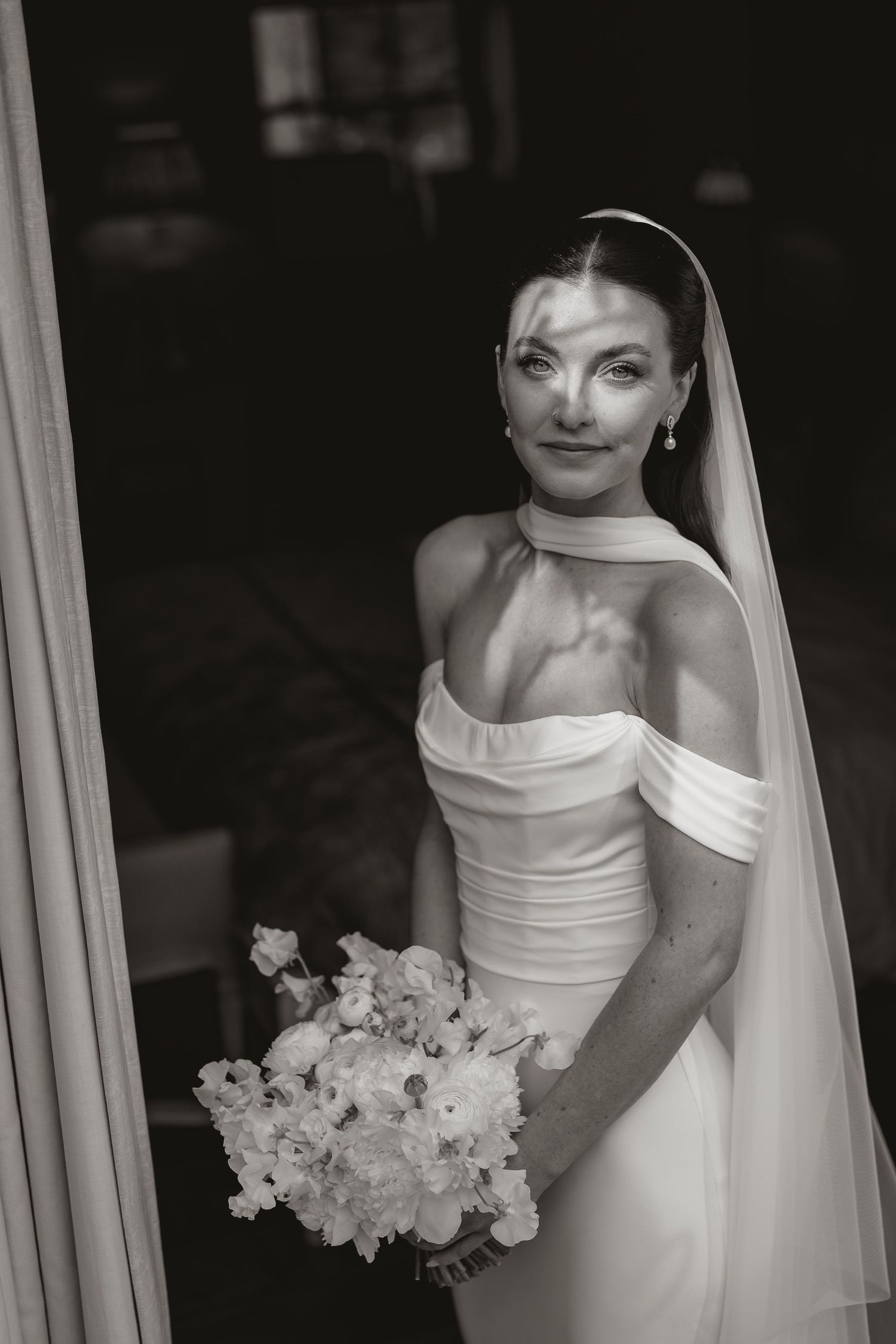 Black and white portrait of a bride in an off-shoulder gown holding a floral bouquet, wearing a veil and choker necklace.