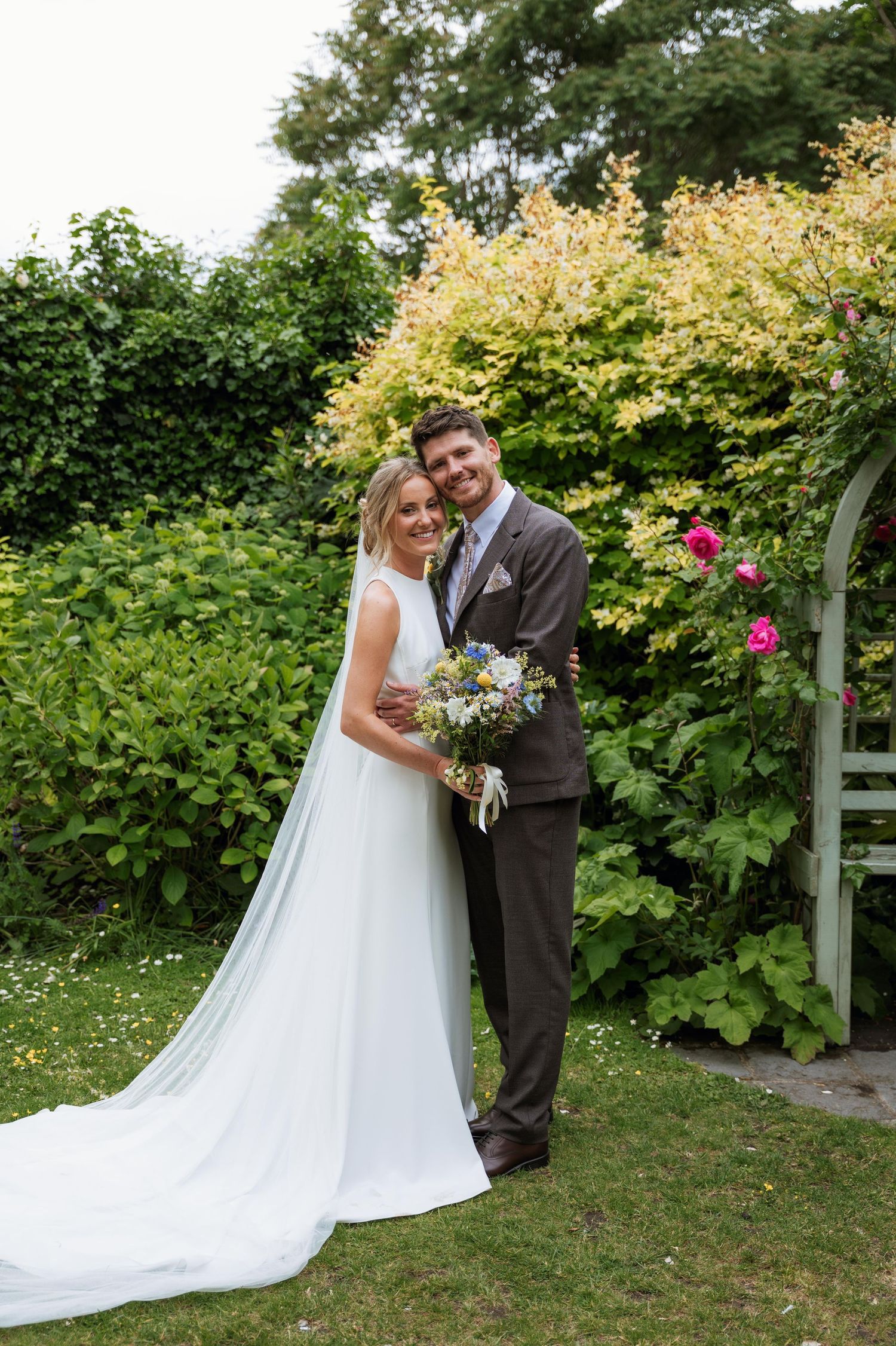 A couple in wedding attire embraces in a lush garden setting with flowering bushes and greenery in the background.