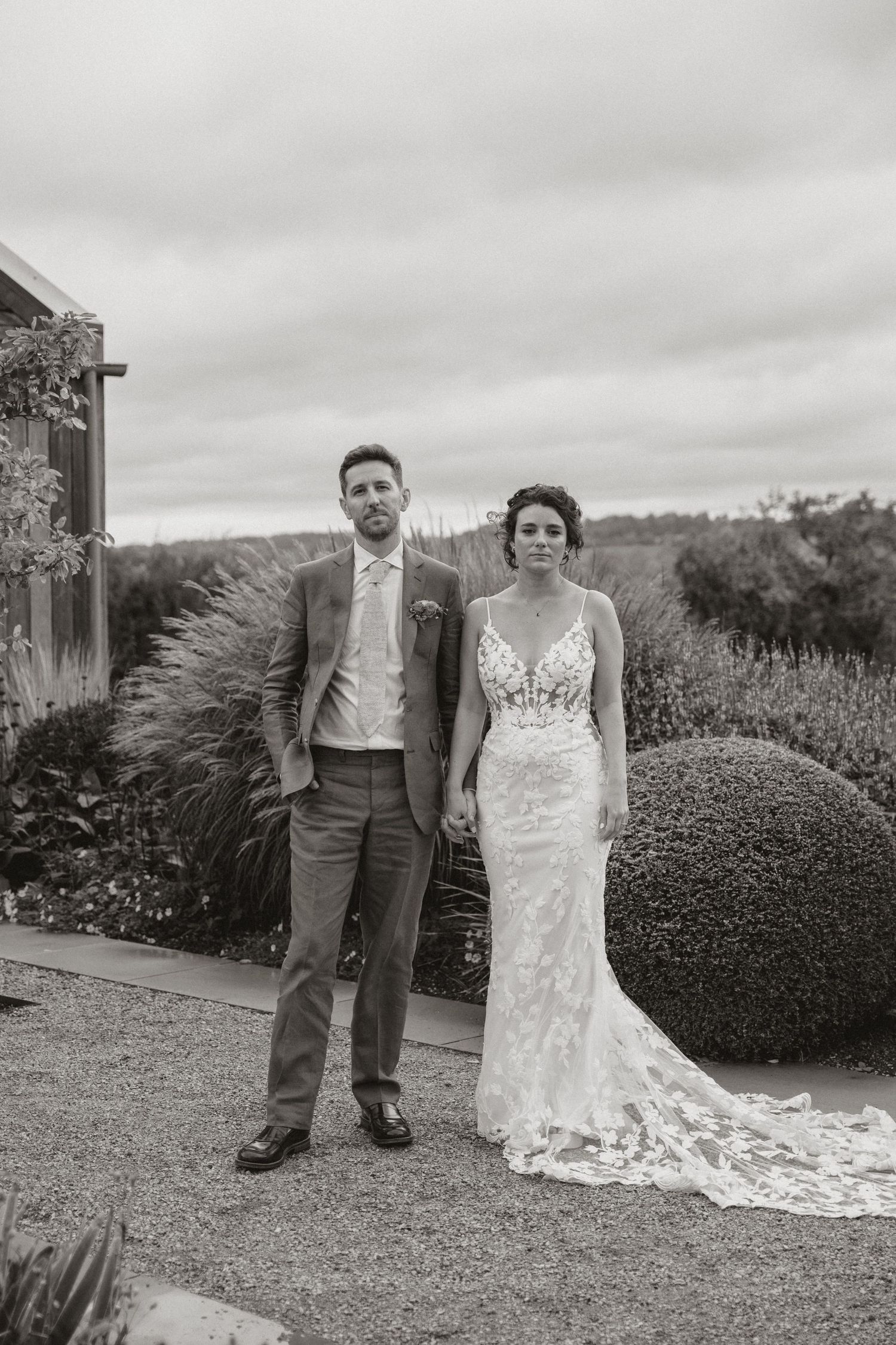 A couple poses together in a garden setting on their wedding day with overcast skies in the background.