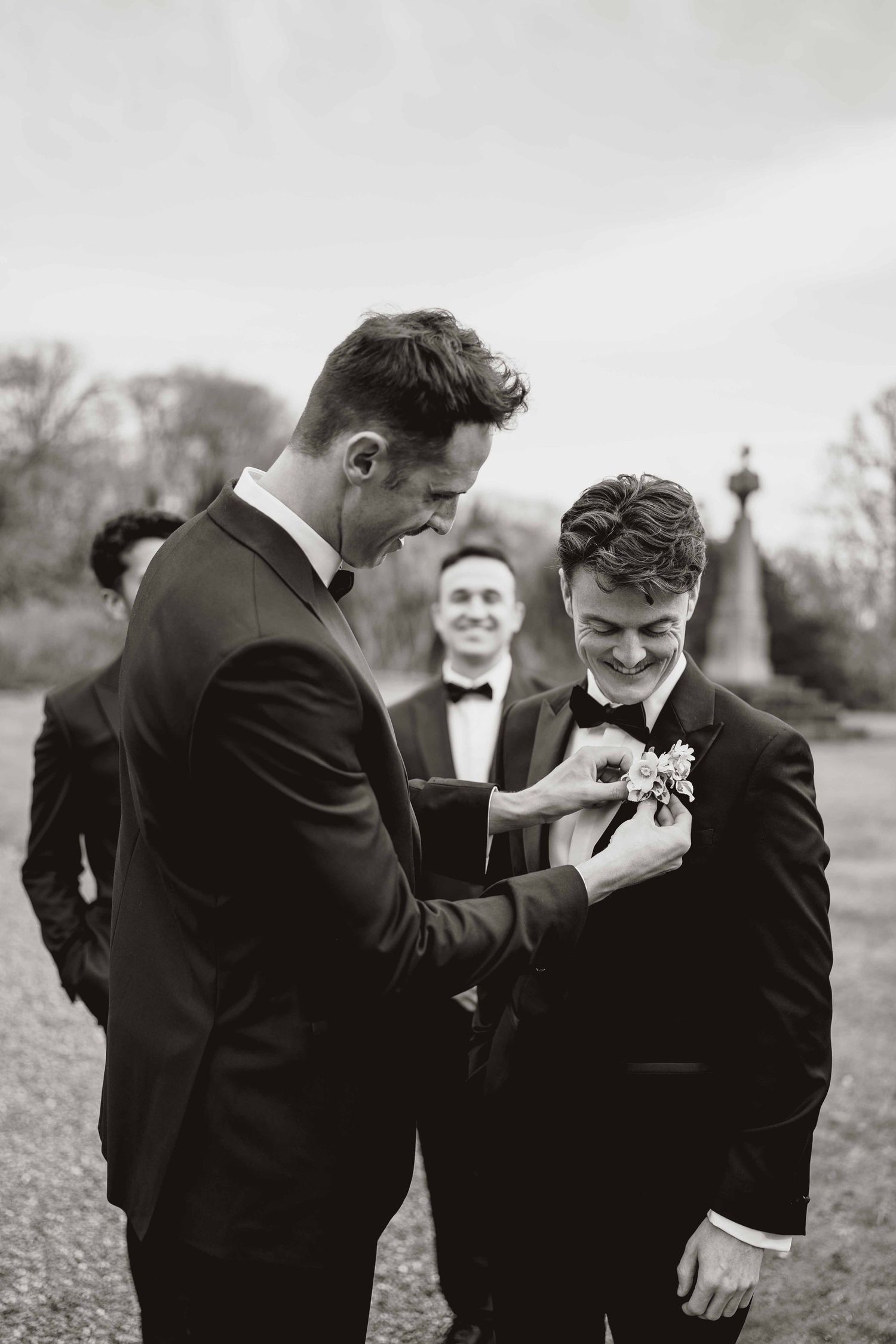 Groomsmen help adjust boutonnieres and bow ties while sharing laughs during outdoor wedding preparations.