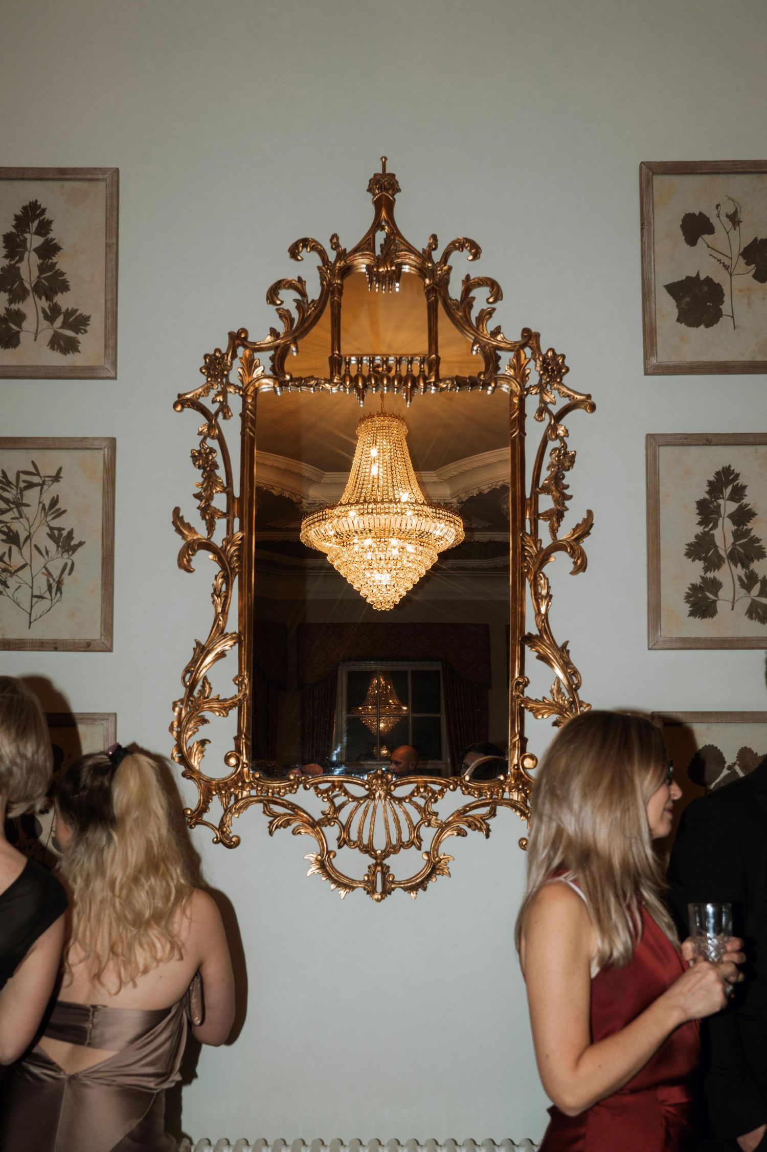 Ornate gold-framed mirror reflects elegant crystal chandelier on wall decorated with botanical prints at formal gathering.