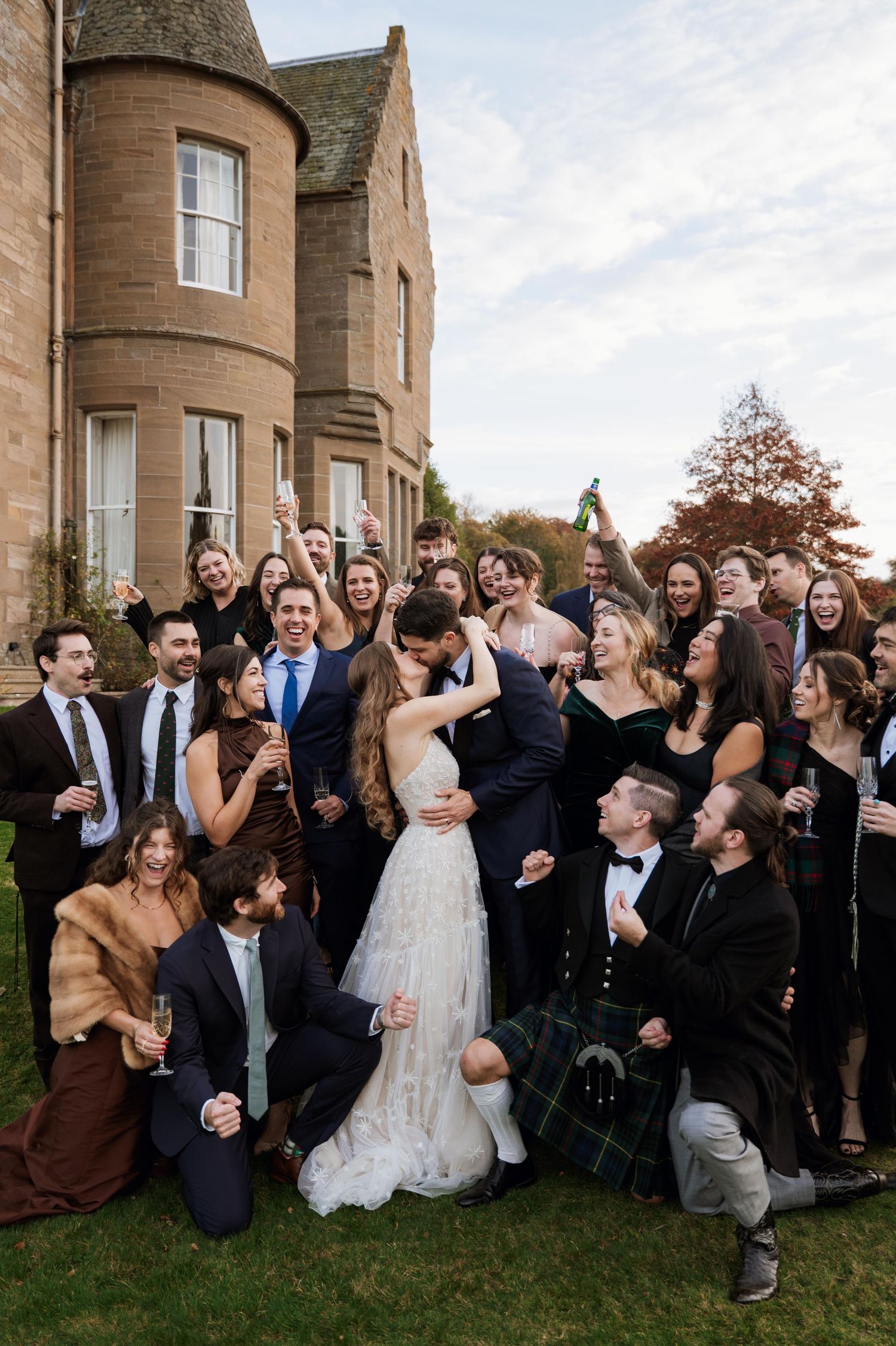 Large wedding party celebrates joyfully outside stone castle venue during autumn celebration.
