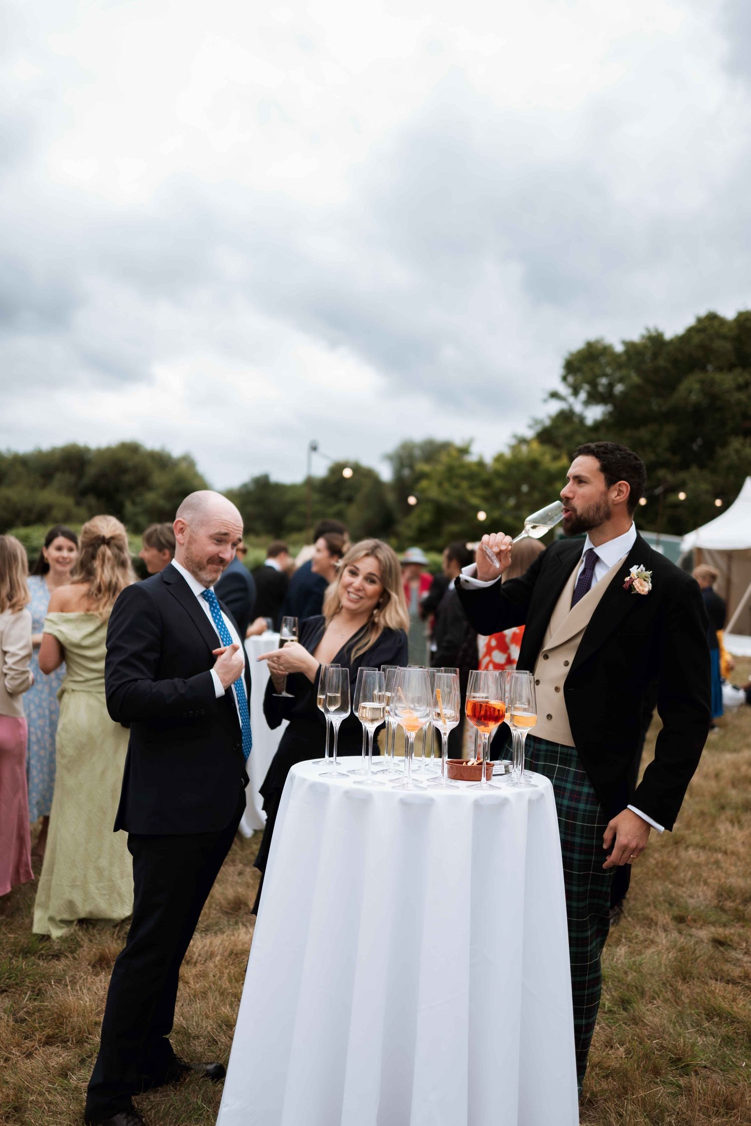 Guests gather around a cocktail table at an outdoor wedding reception with champagne glasses under cloudy skies.